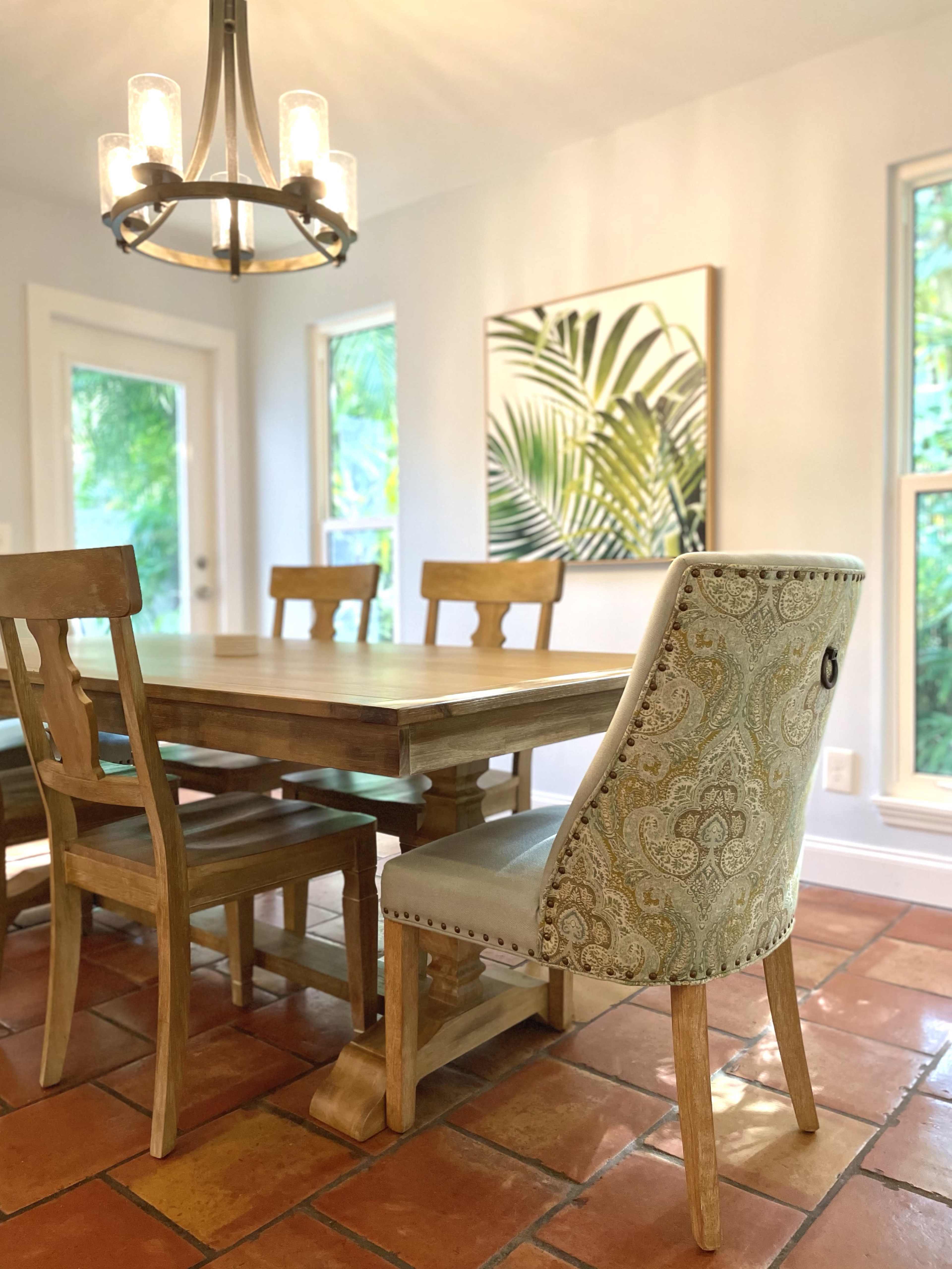 A wooden dining table with six chairs is positioned under a chandelier in a room with large windows and a tropical-themed wall art.