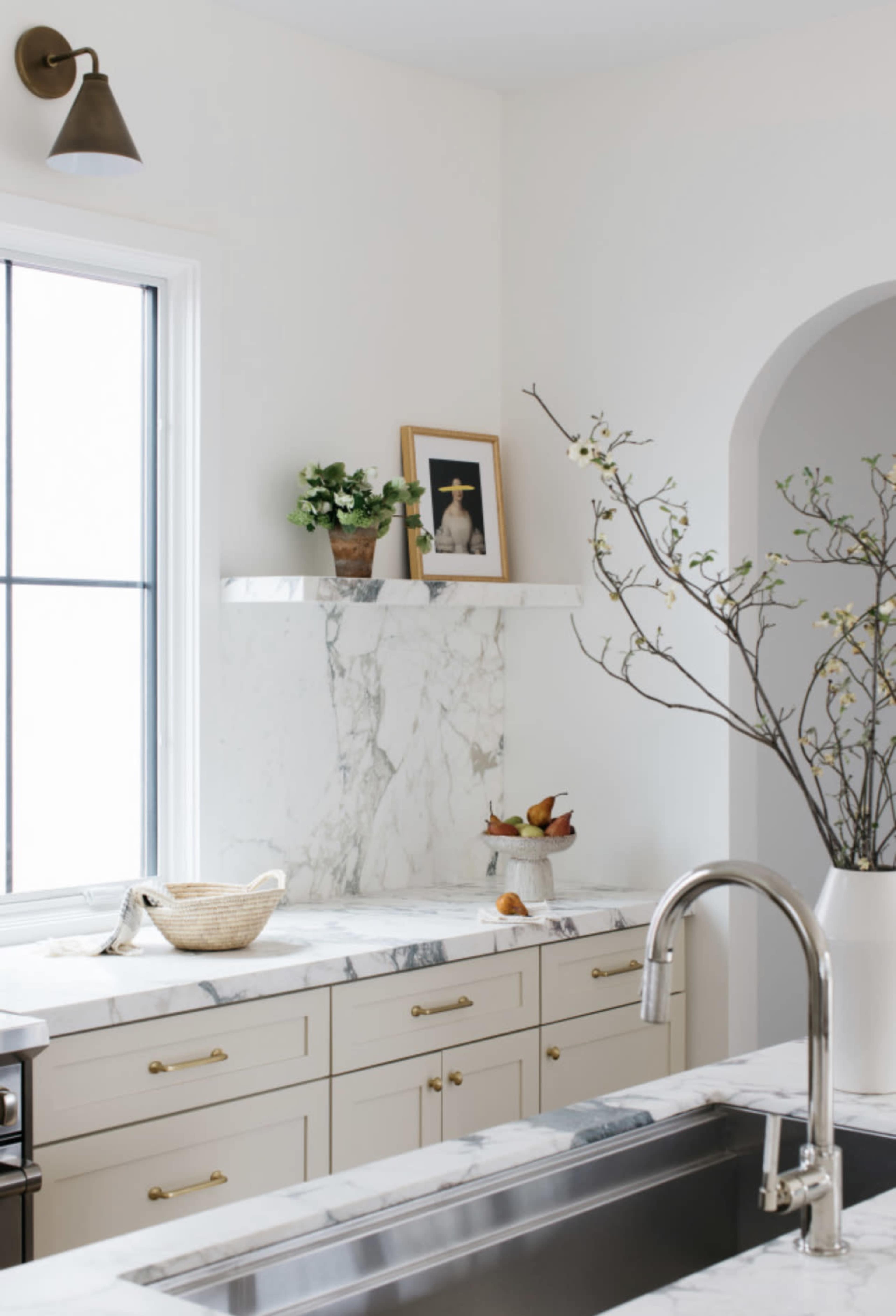 The image shows a modern kitchen with marble countertops, a sink, and a window providing natural light, featuring a vase with branches and a framed photo on the wall.