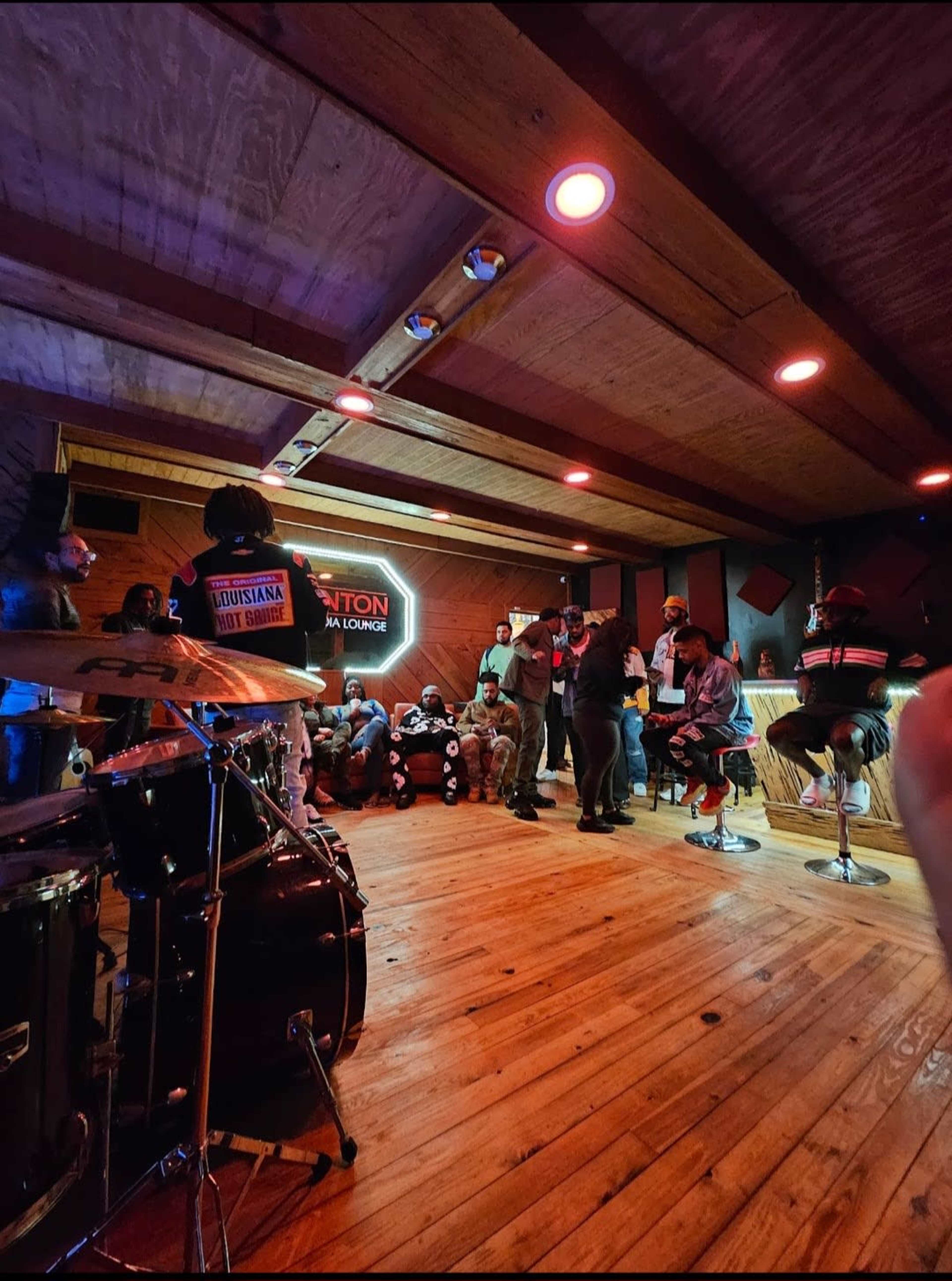 A group of people gathers in a dimly lit lounge, with a drum set visible in the foreground and a neon sign on the wall.
