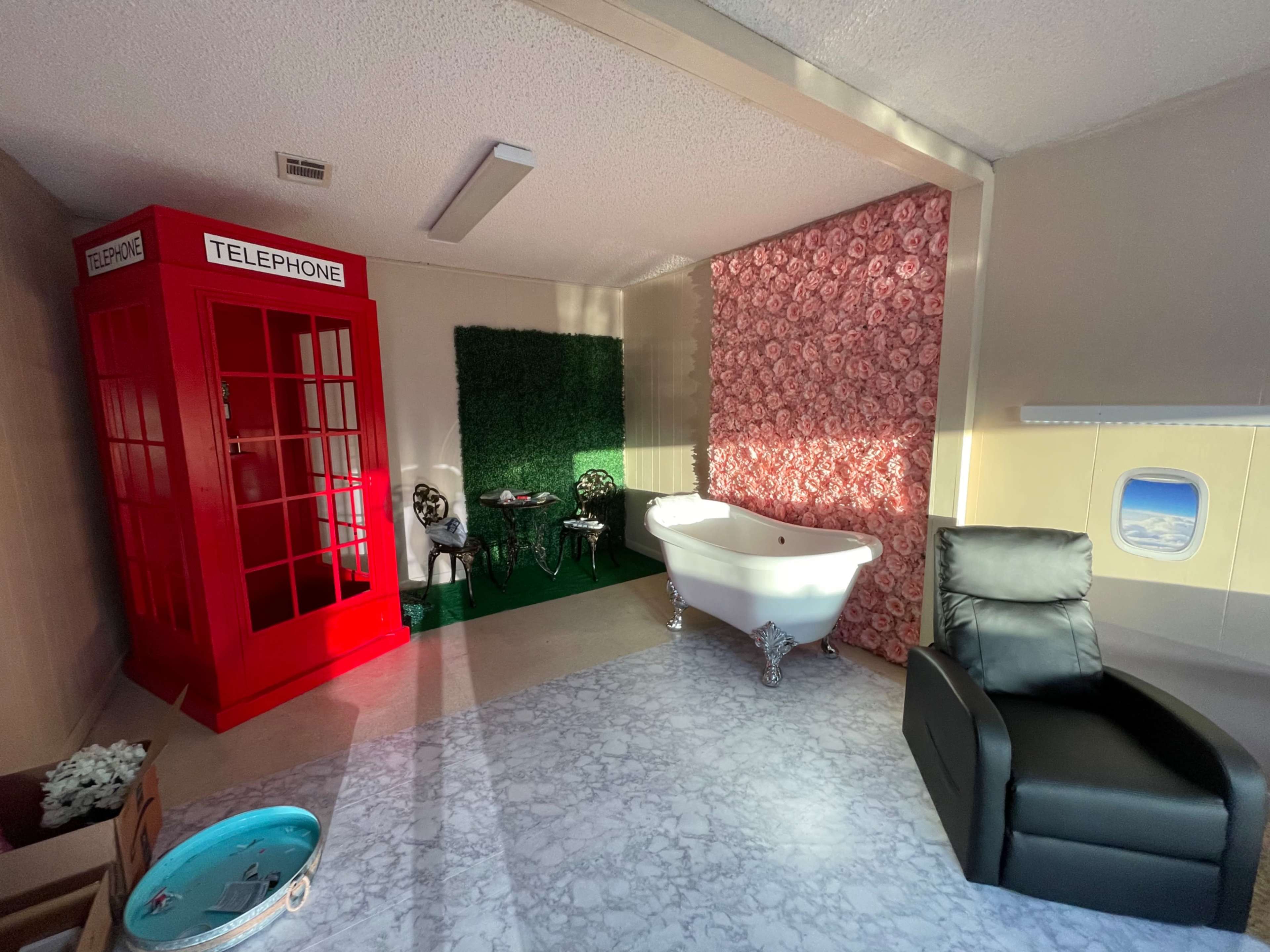 The image shows a small interior space featuring a red telephone booth, a green wall, a vintage bathtub, and a black recliner chair.