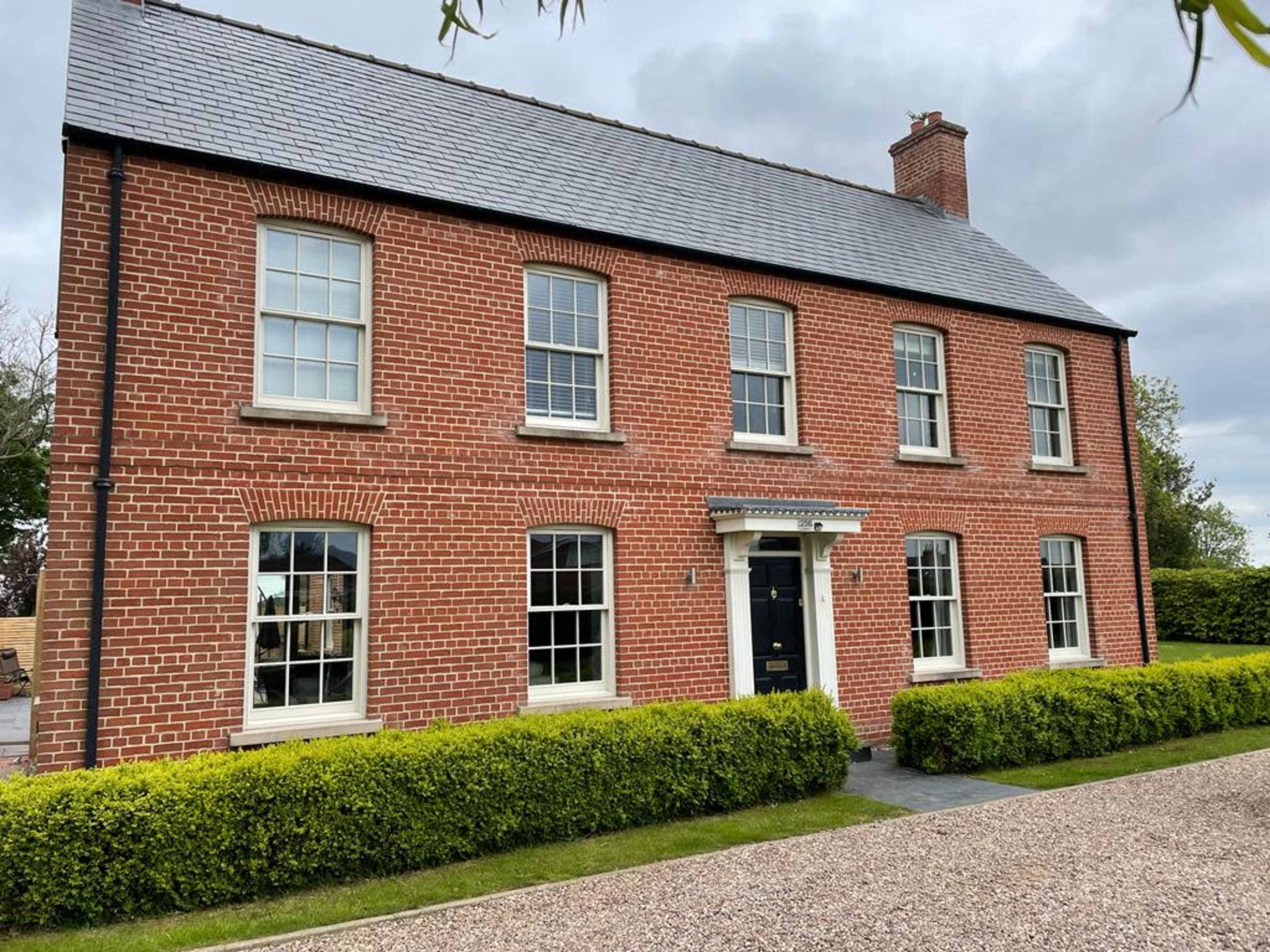 A red-brick house with white windows and a black front door is set against a gray sky and surrounded by neatly trimmed hedges.
