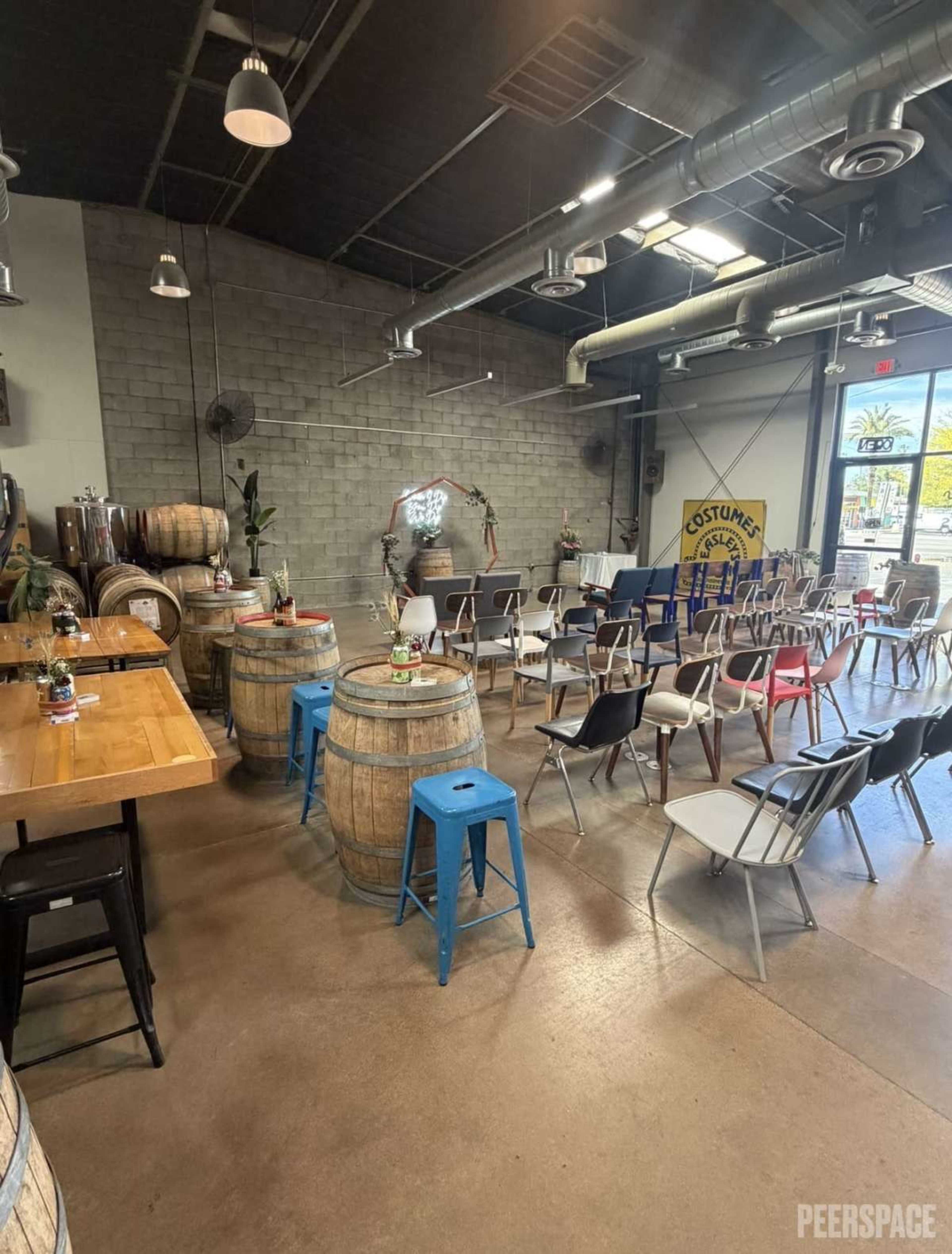 The image shows a spacious venue filled with wooden barrels, metal chairs, and a long table, illuminated by natural light through large windows.