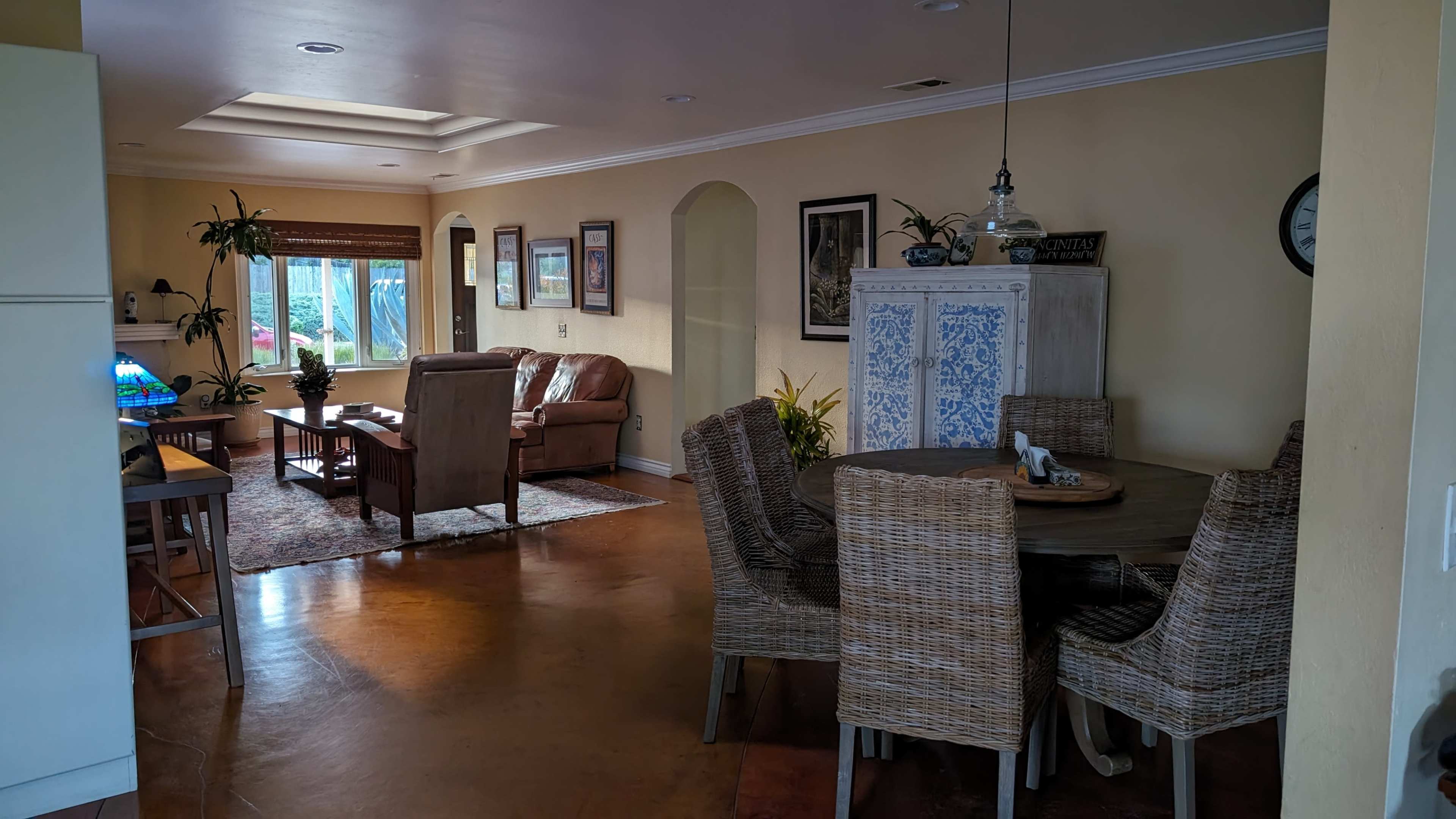 The image shows a living and dining area with a table set for six, a brown leather sofa, and various decorative elements on the walls.
