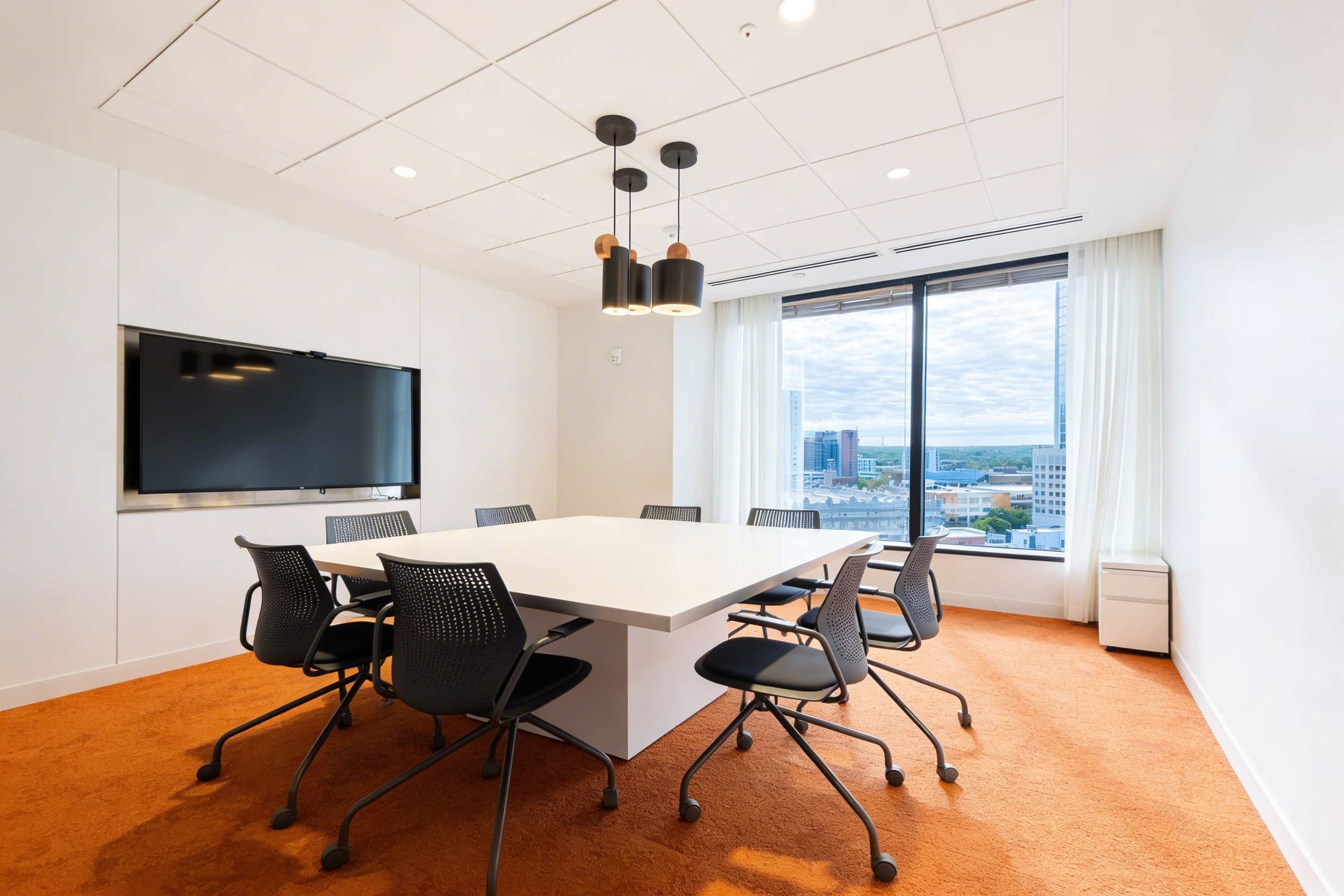 A modern conference room features a large white table surrounded by black mesh chairs, a wall-mounted TV, and a window offering a view of the cityscape.