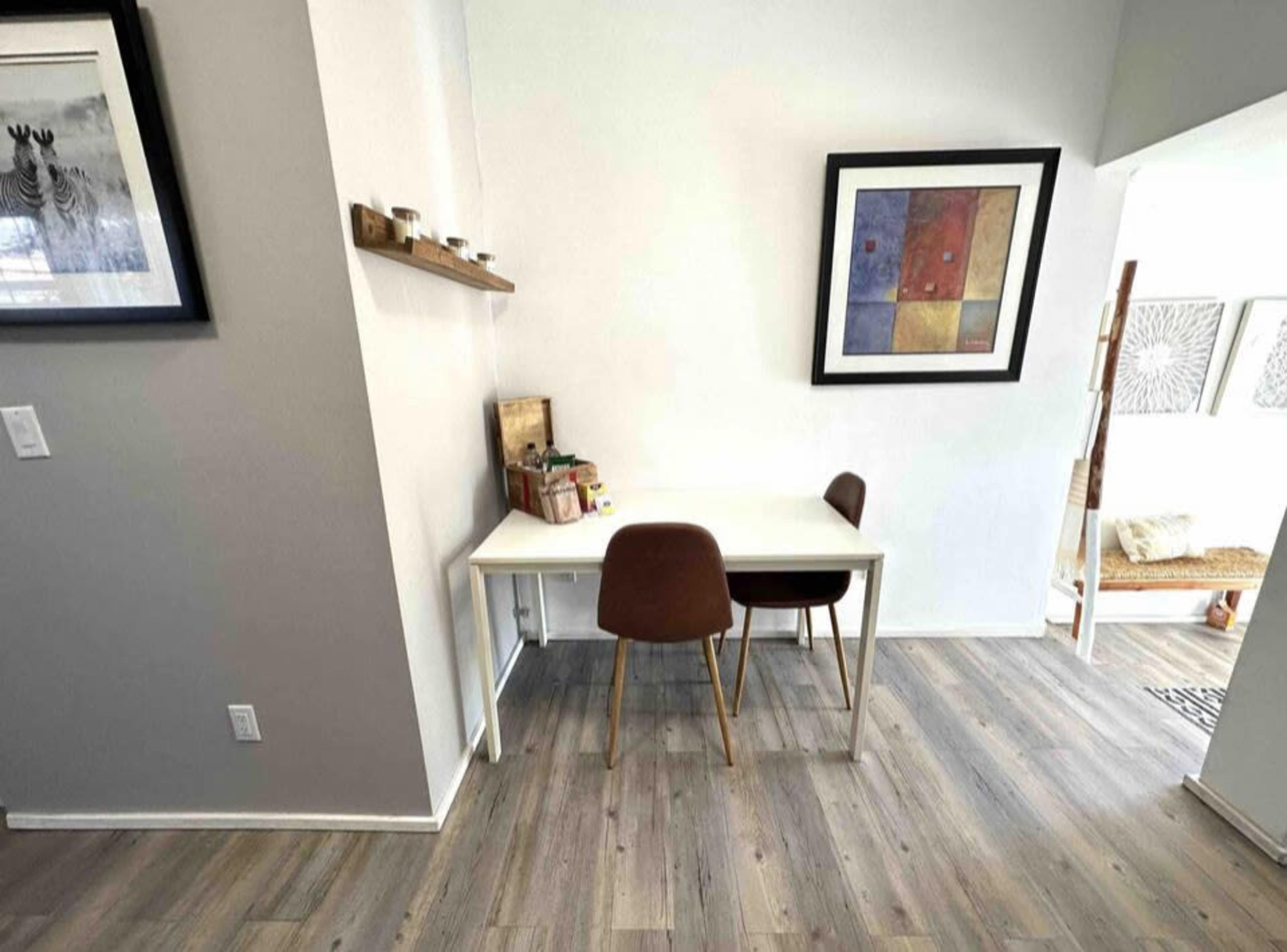 A small dining area features a white table with two chairs against a light gray wall, adorned with a framed abstract artwork and a shelf holding small items.