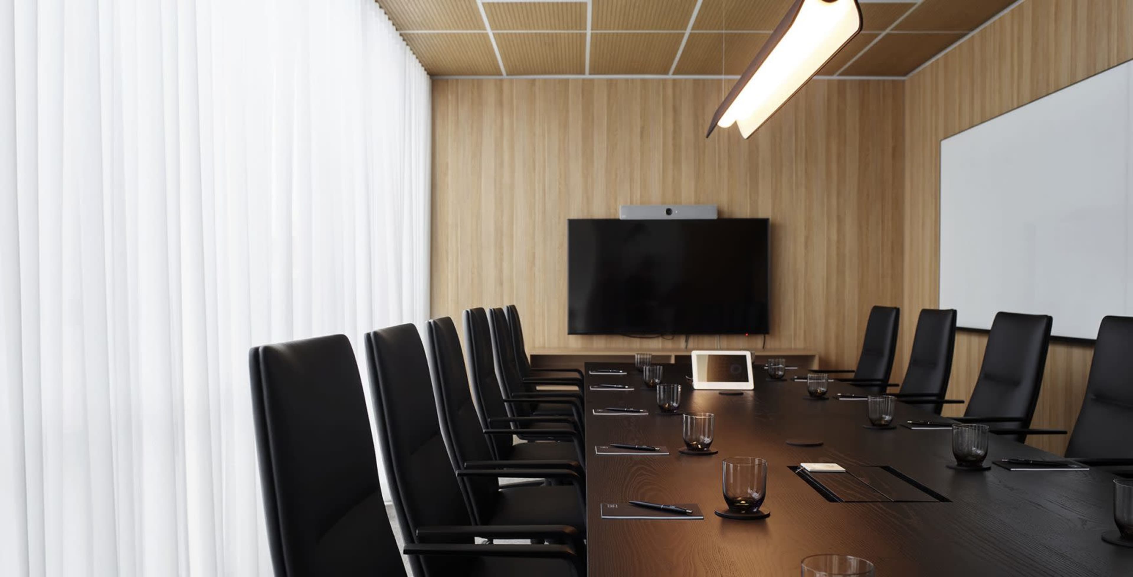 A conference room features a long table with black chairs, a large screen on the wall, and natural light filtering through sheer curtains.