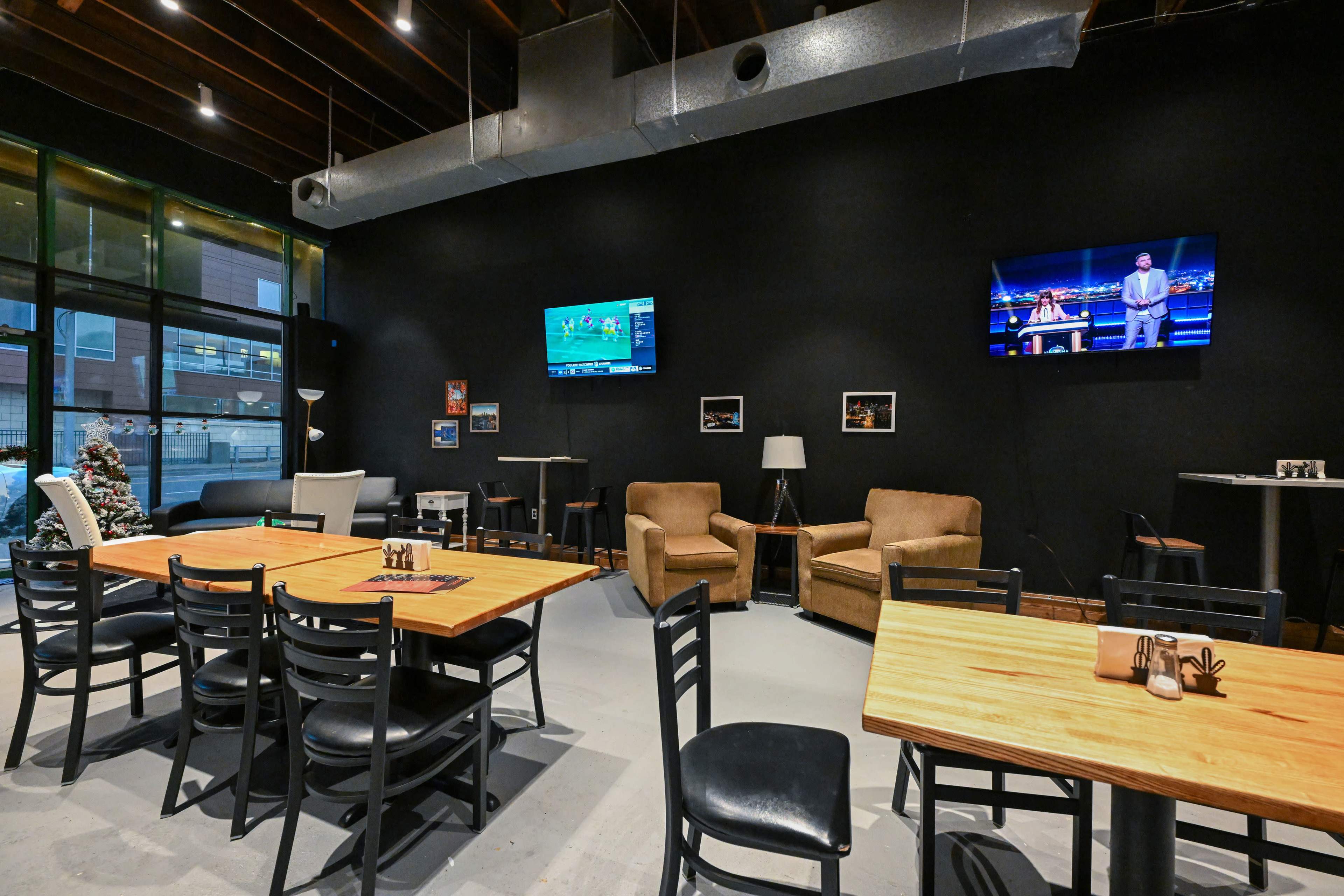 The image features a modern restaurant interior with black walls, wooden tables, and seating, two televisions mounted on the wall, and a Christmas tree in the corner.
