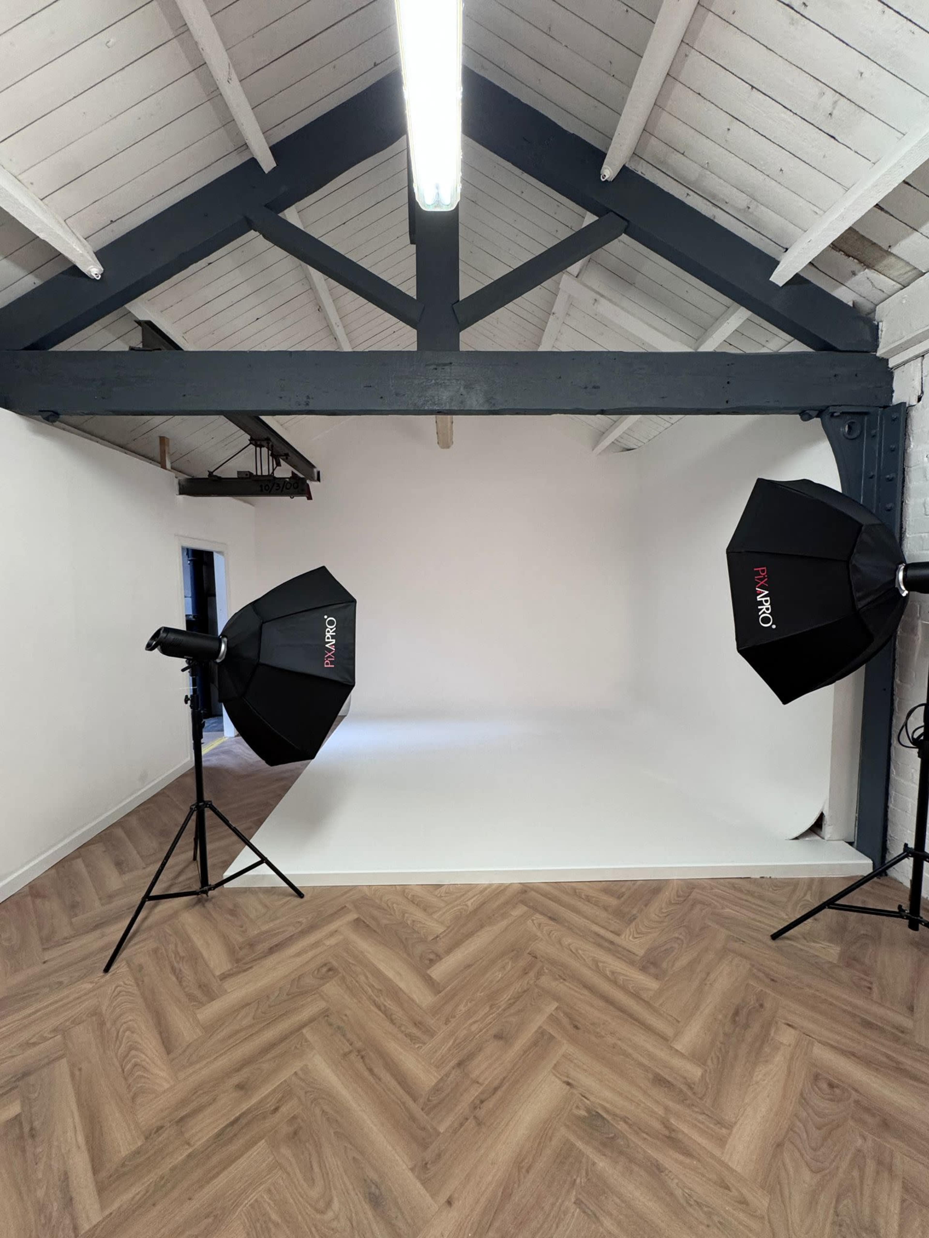 The image shows a spacious photography studio with two lighting umbrellas set up facing a white backdrop.