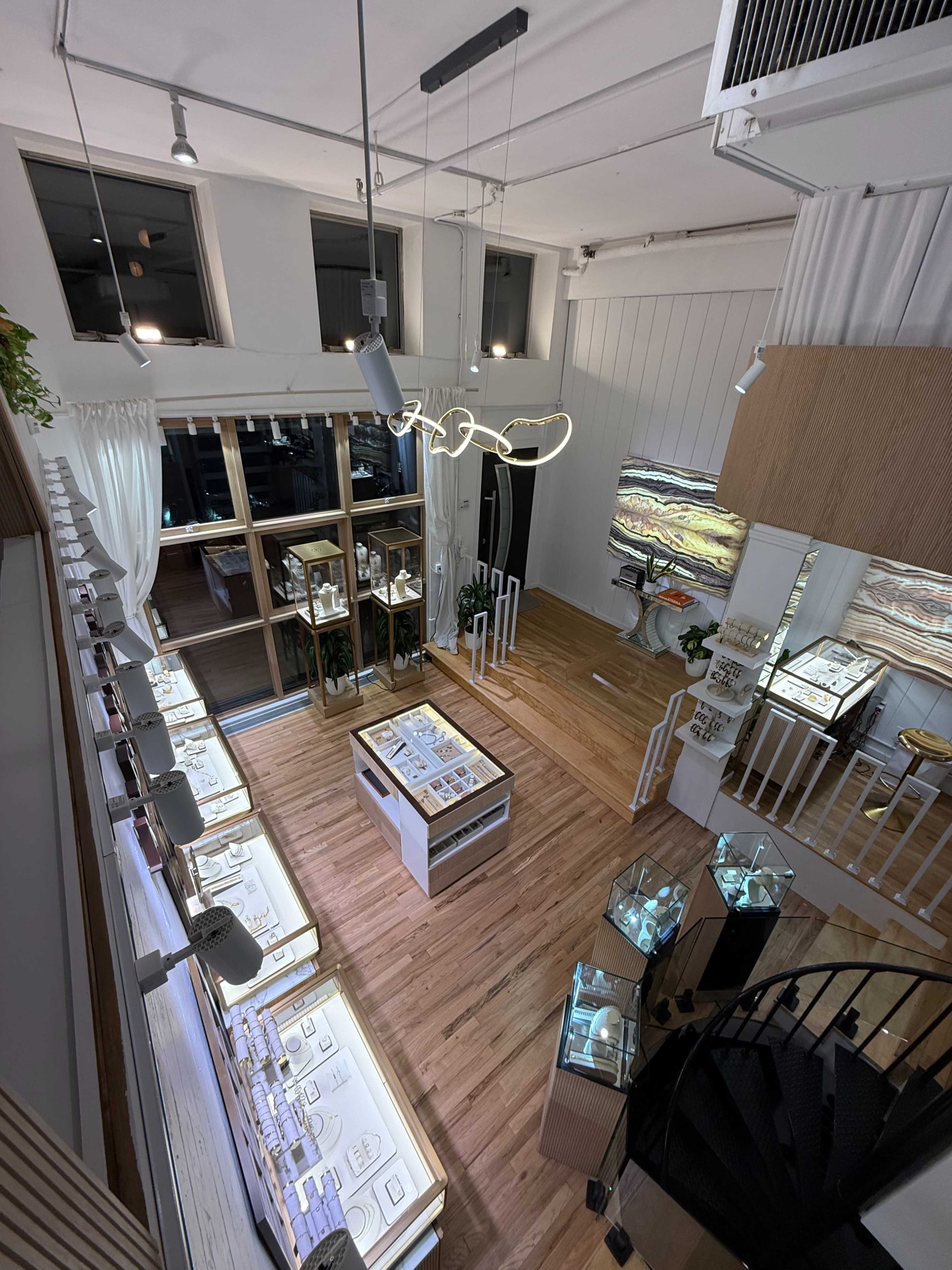 The image shows a bright jewelry store with multiple display cases, wooden flooring, and a spiral staircase, featuring large windows that allow natural light to flood the space.