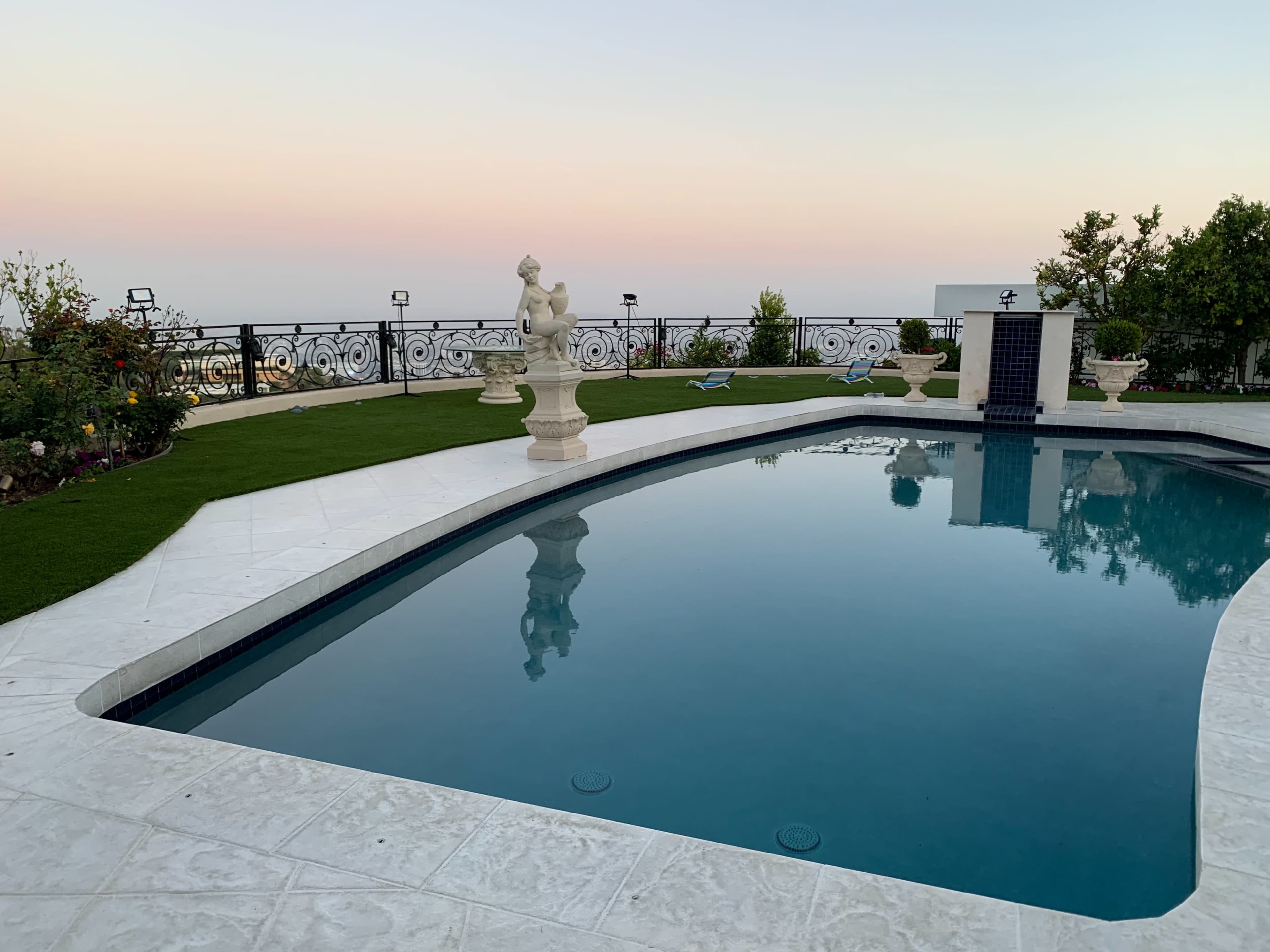The image shows a pool surrounded by a tiled patio, featuring a sculpture and landscaped gardens against a pastel sky.