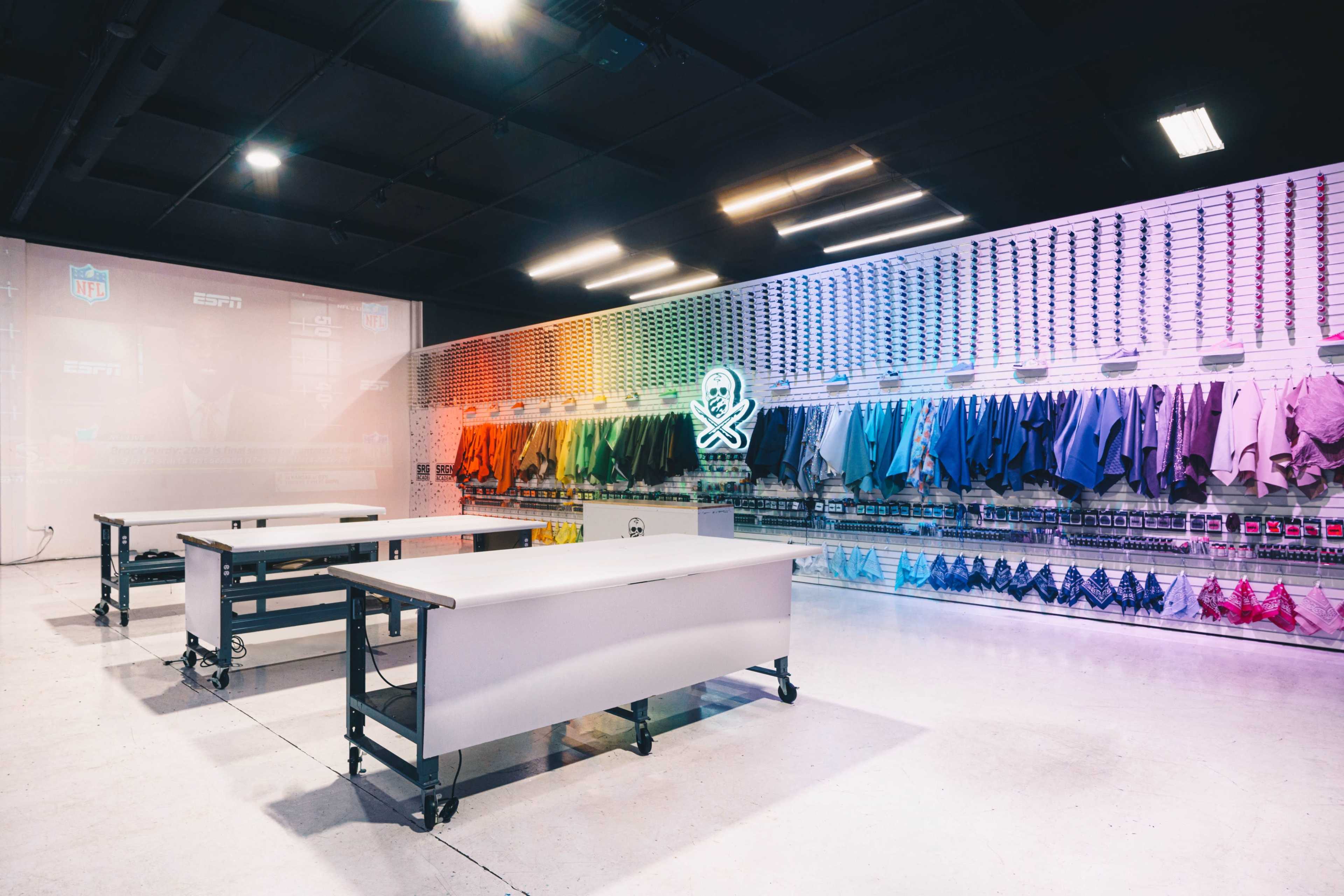 The image shows a modern workspace with several tables and a wall displaying a variety of colorful clothing items, organized by color, alongside sports branding elements.
