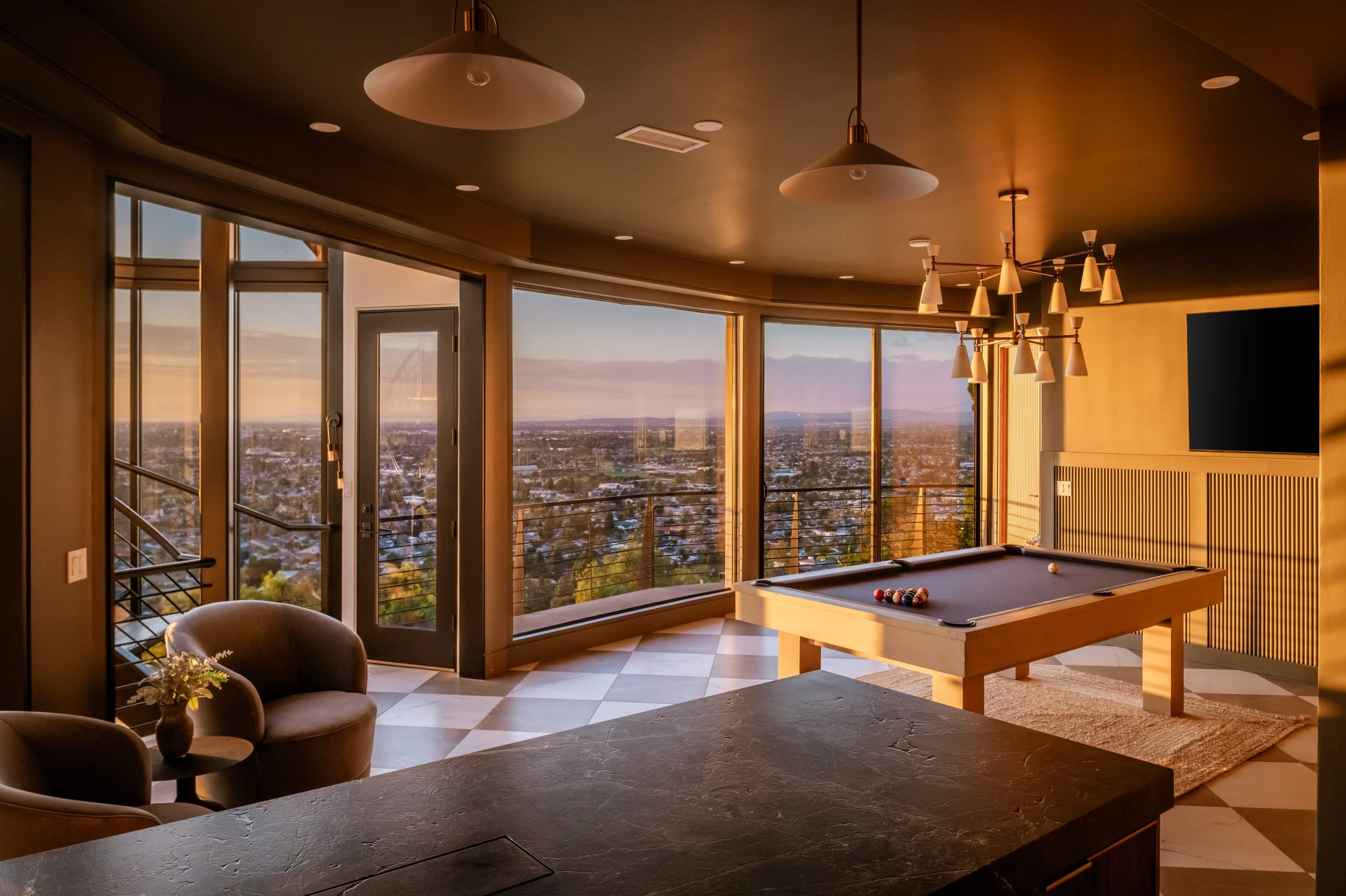 A modern game room features a billiards table and large windows providing a view of the city skyline at sunset.
