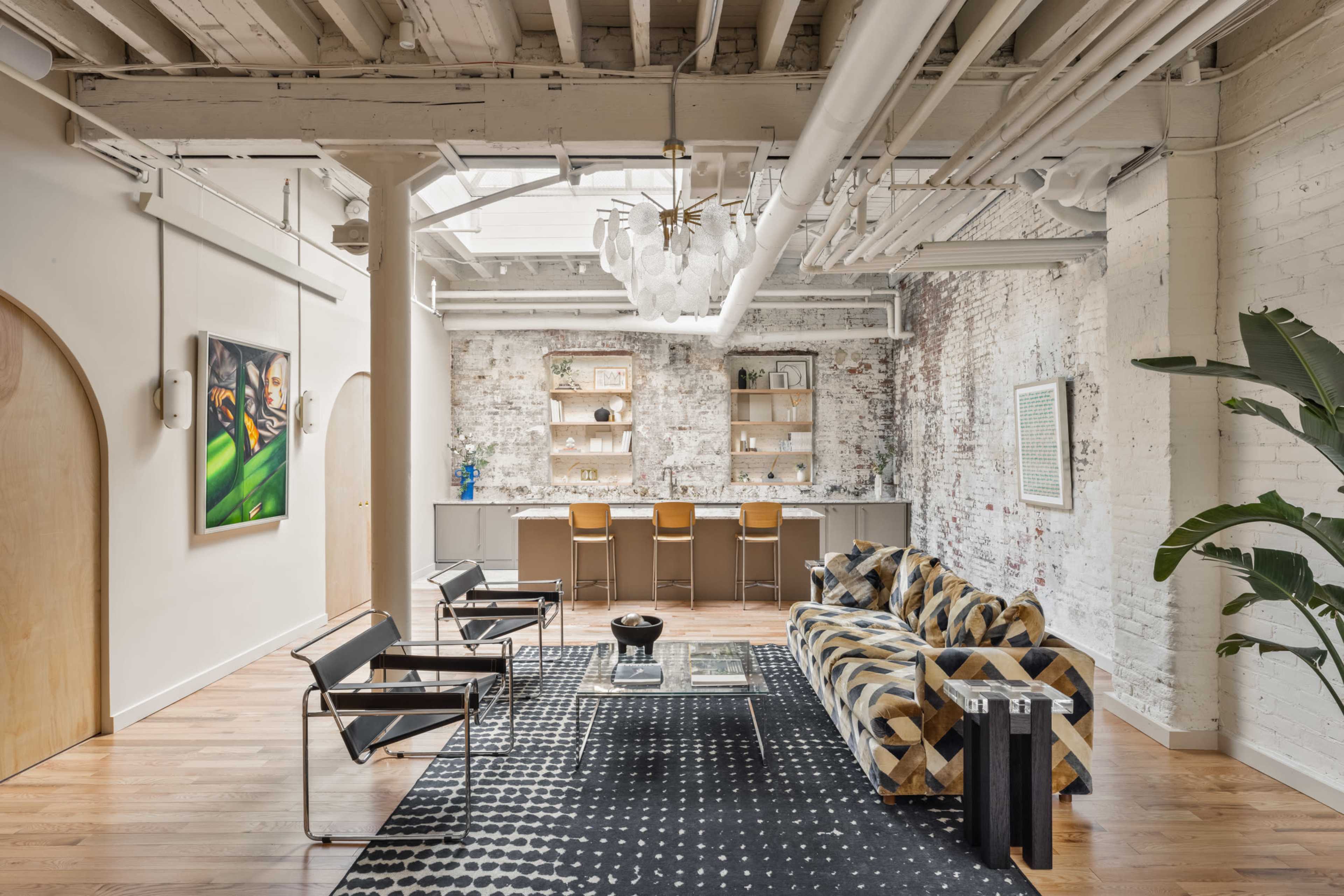 The image depicts a spacious, modern living area with exposed brick walls, wooden flooring, a patterned sofa, and a bar setup in the background.