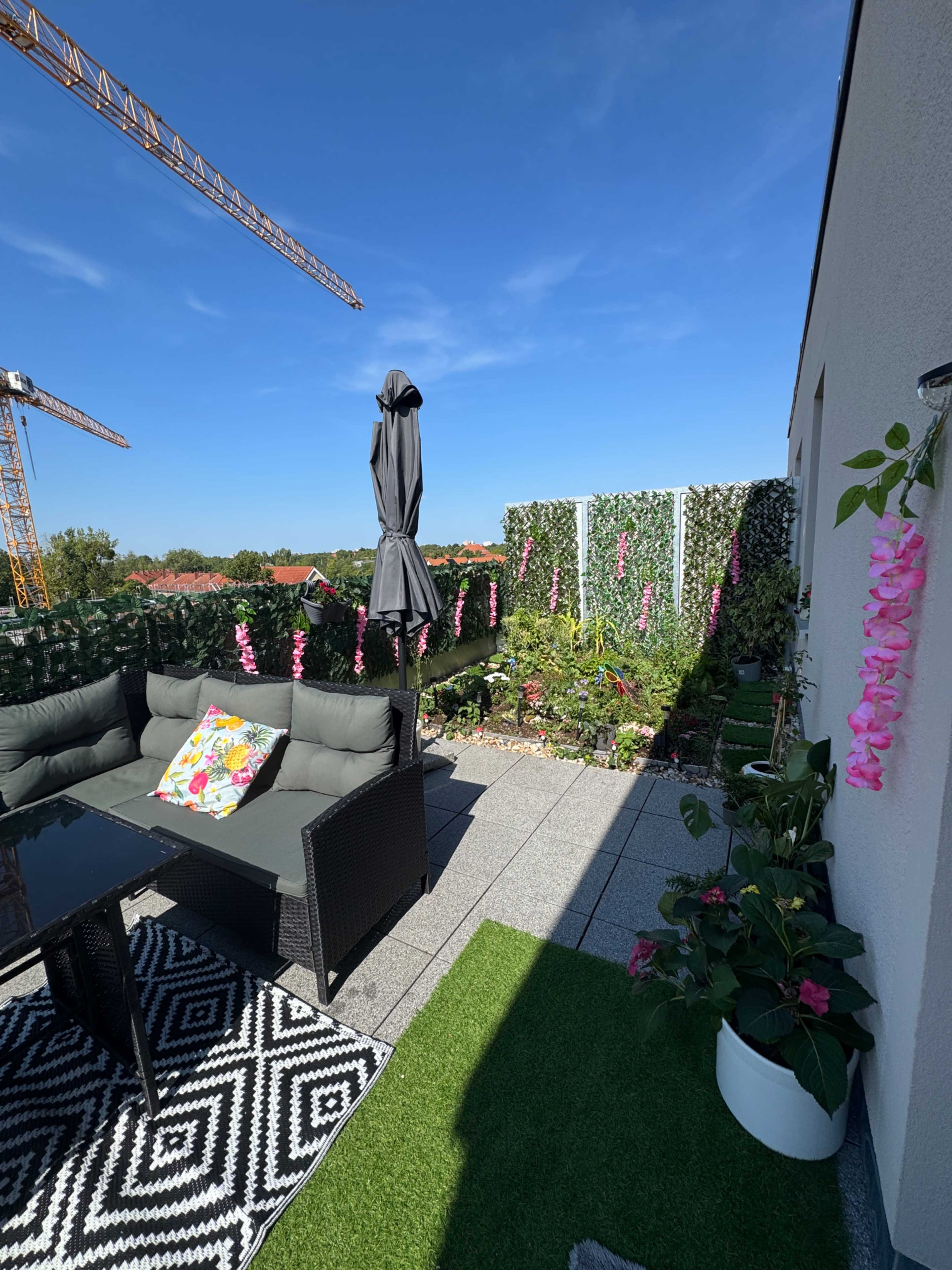 The image shows a rooftop terrace featuring a black outdoor sectional sofa, a table, and lush greenery adorned with pink flowers, along with construction cranes visible in the background under a clear blue sky.