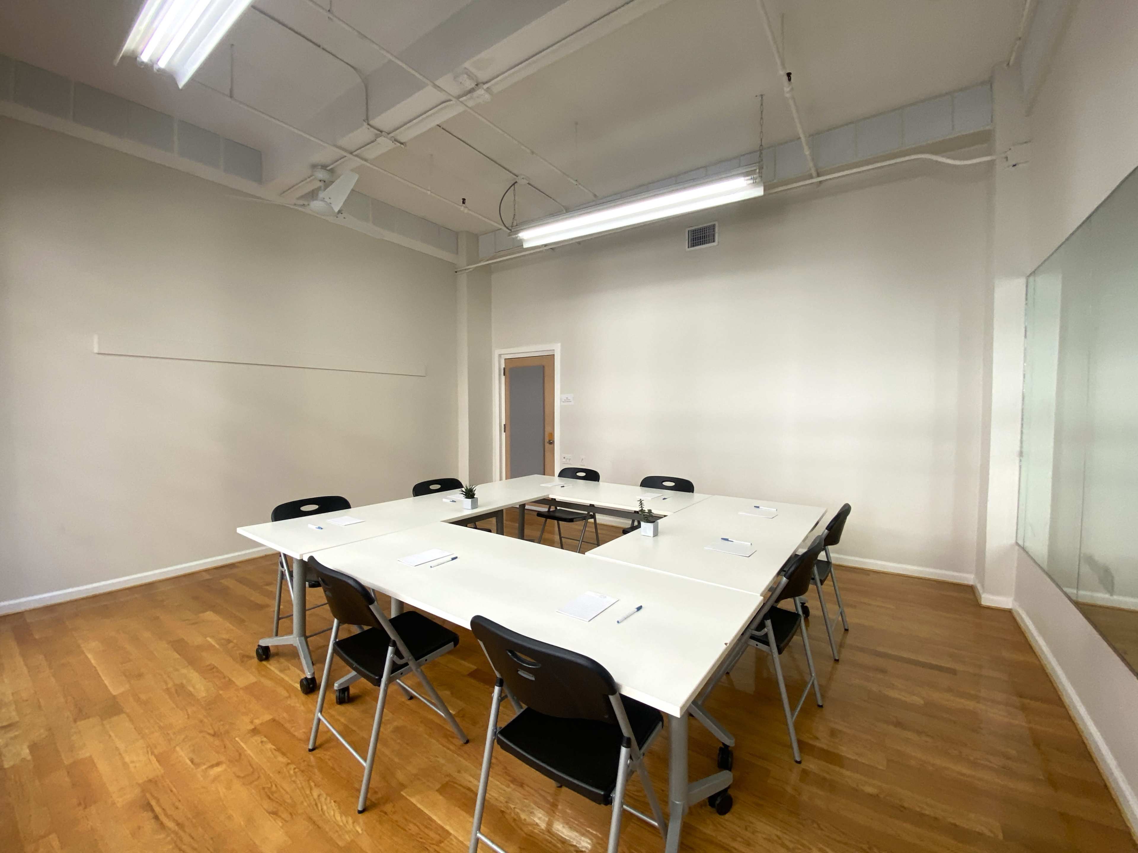A meeting room features a large table surrounded by chairs, with a plain wall and a mirror on one side.