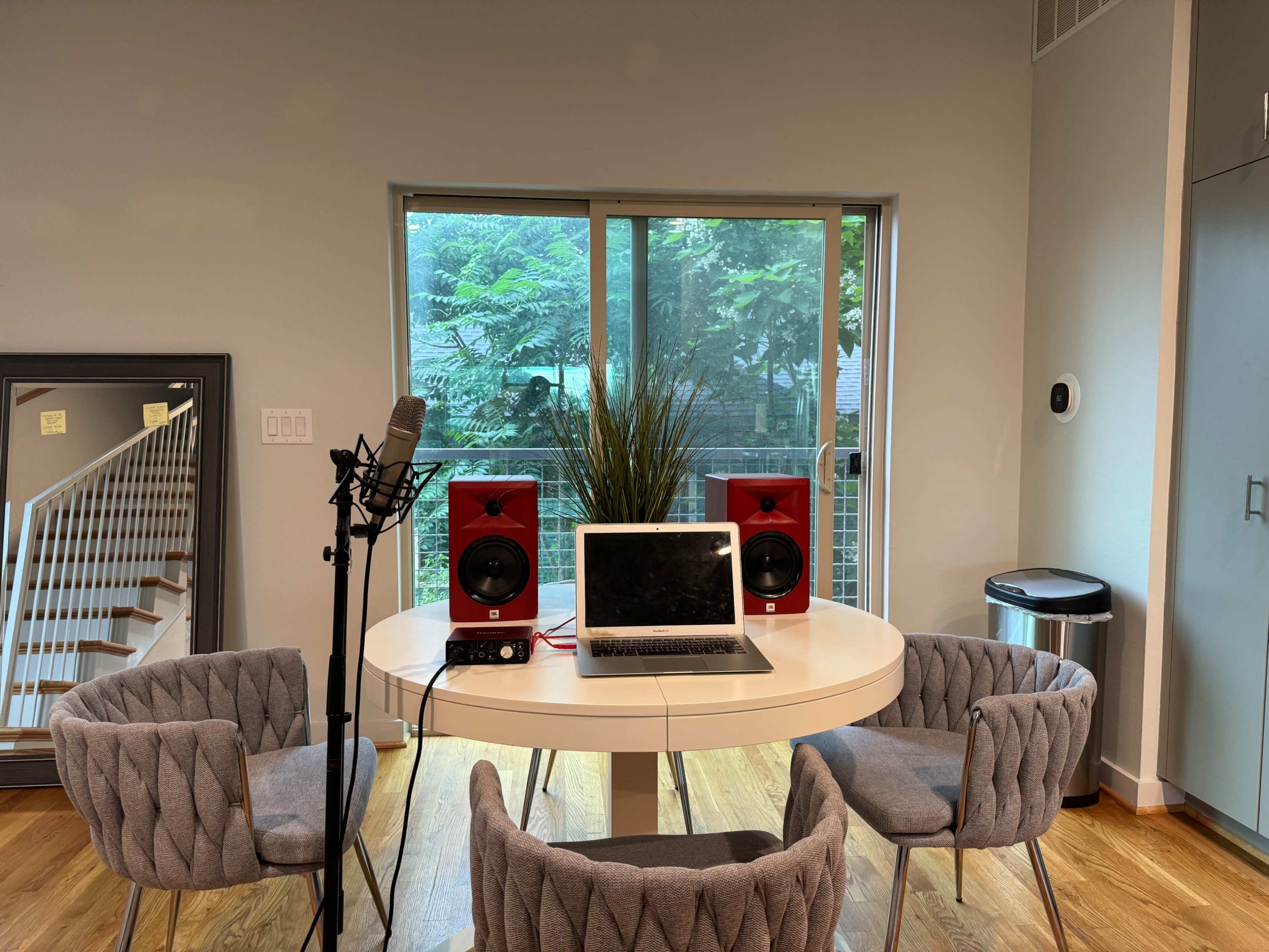 A modern workspace features a round table with a laptop and studio monitors, surrounded by four chairs, next to a large window with greenery outside.
