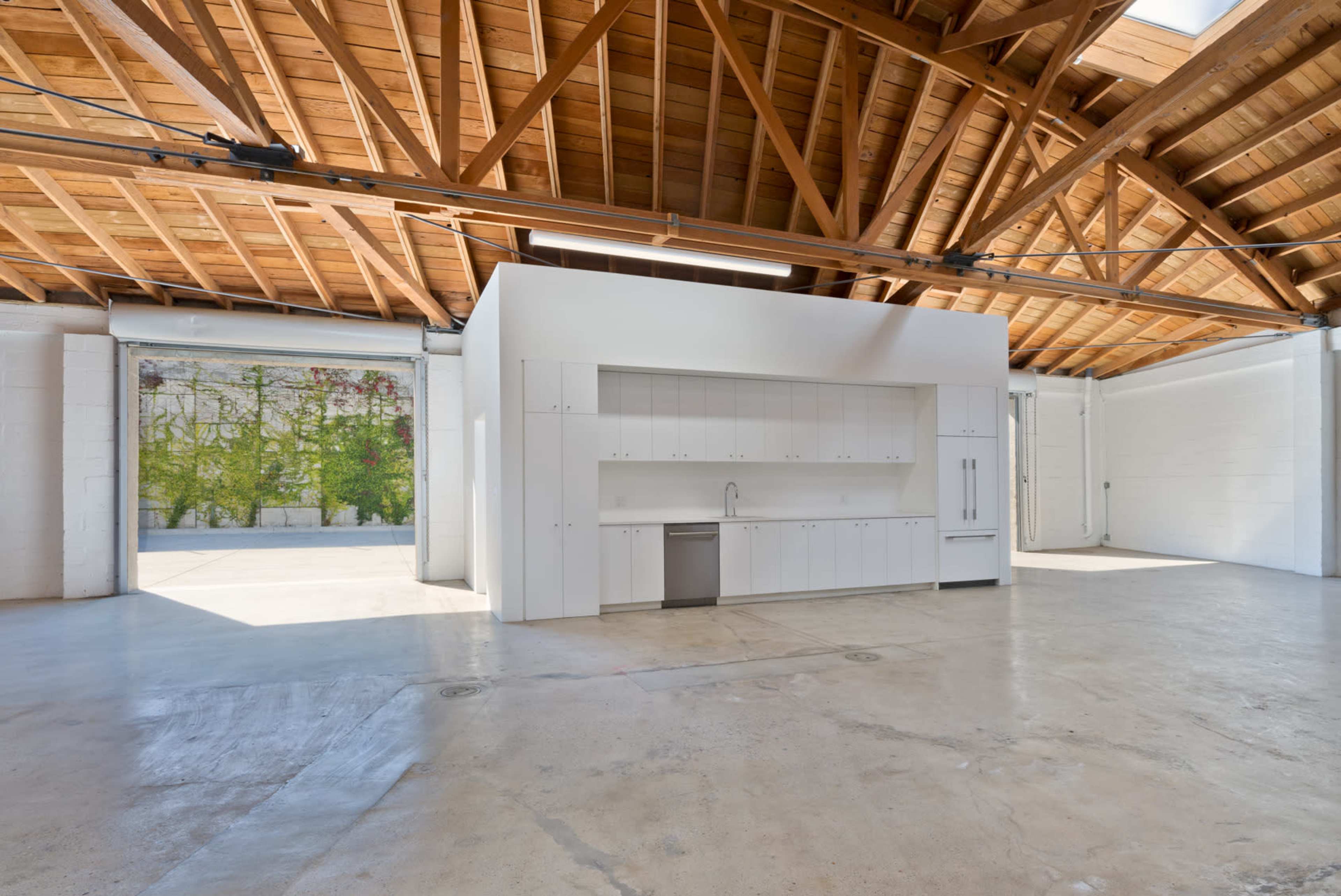 The image shows a spacious, modern kitchen with white cabinetry set in an industrial-style room featuring exposed wooden beams and large windows.