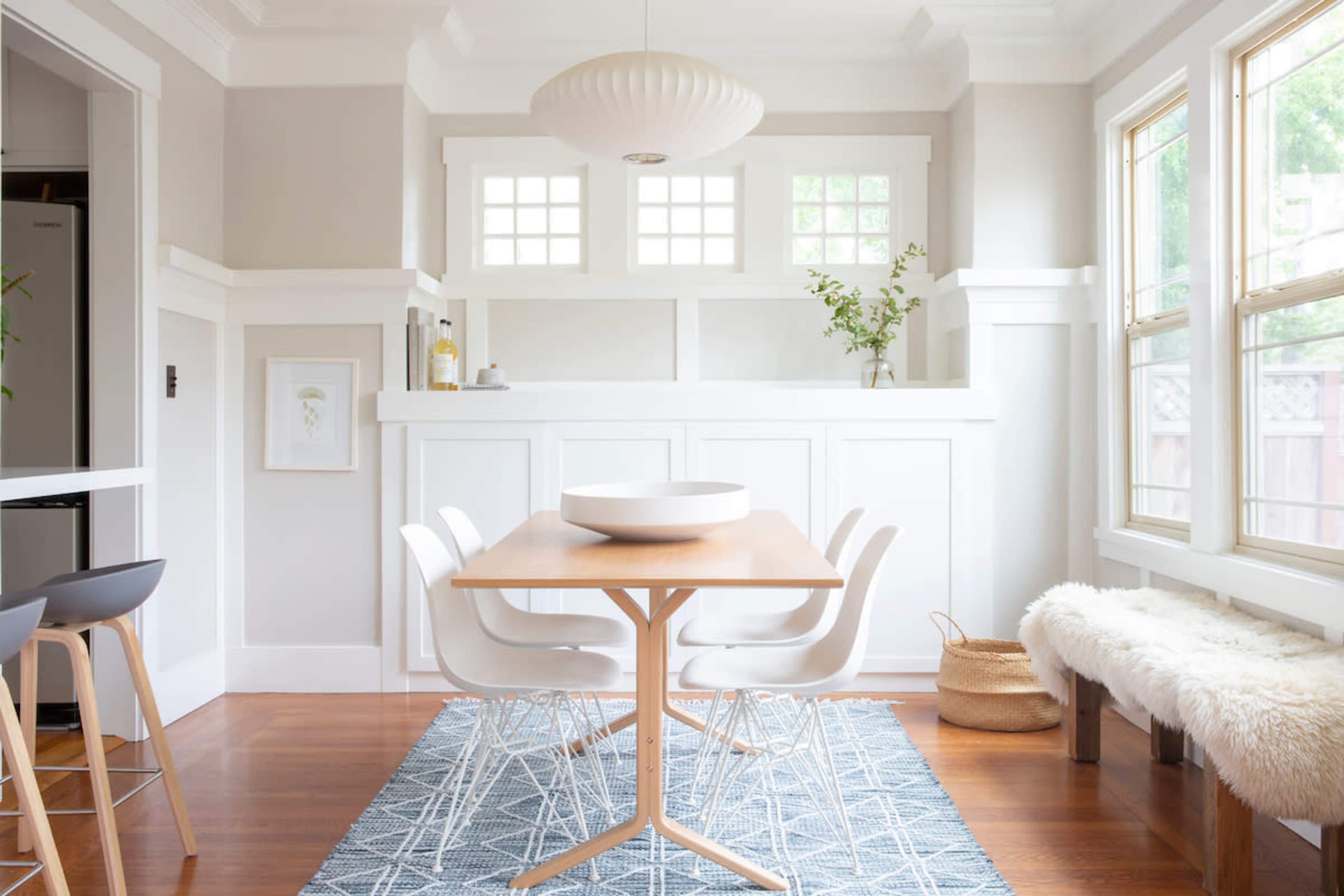 The image shows a modern dining area with a wooden table surrounded by white chairs, set against a backdrop of light-colored walls and large windows.