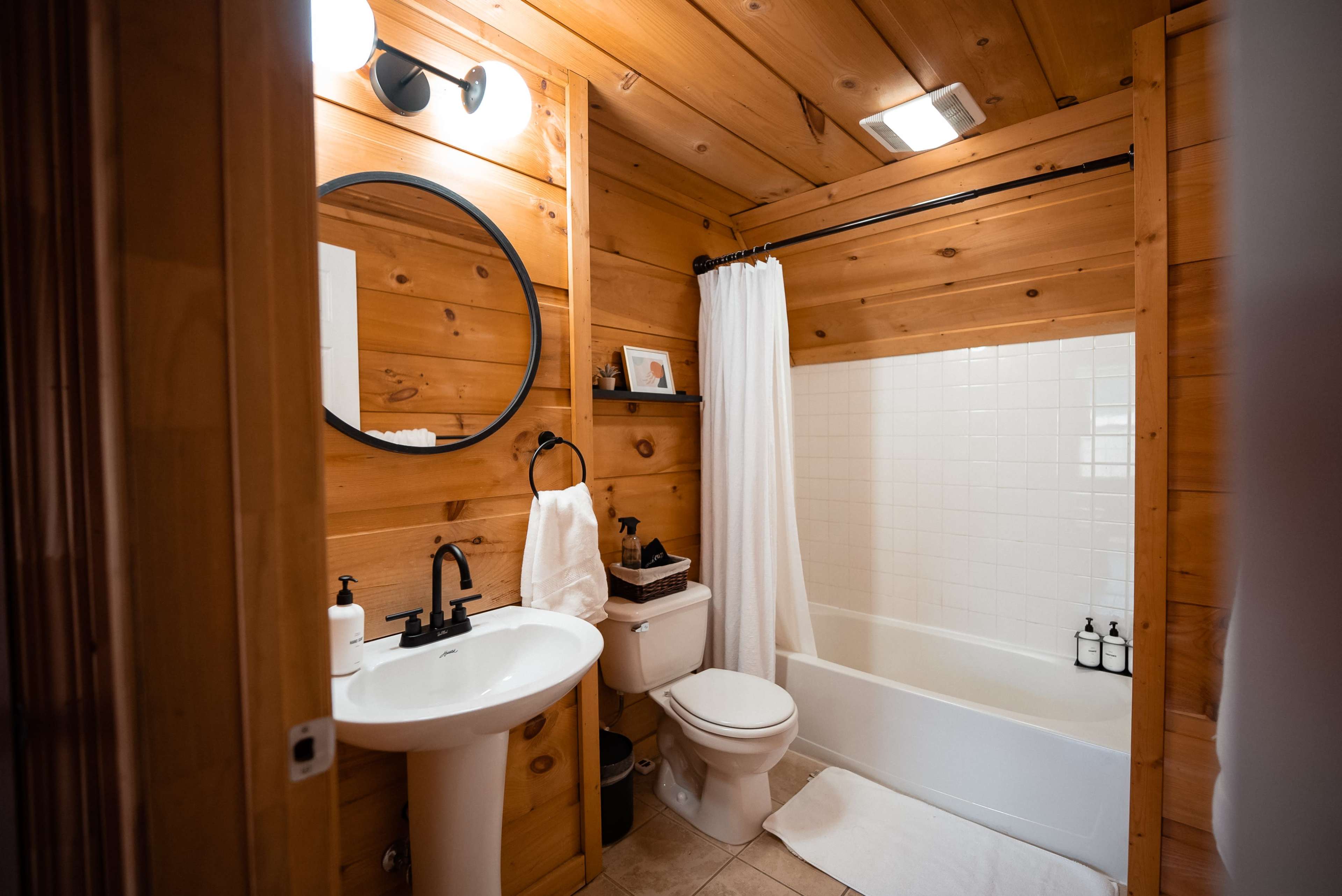 The bathroom features wooden walls, a round mirror above a sink, a white bathtub, and a toilet, with a shower curtain and toiletries on display.