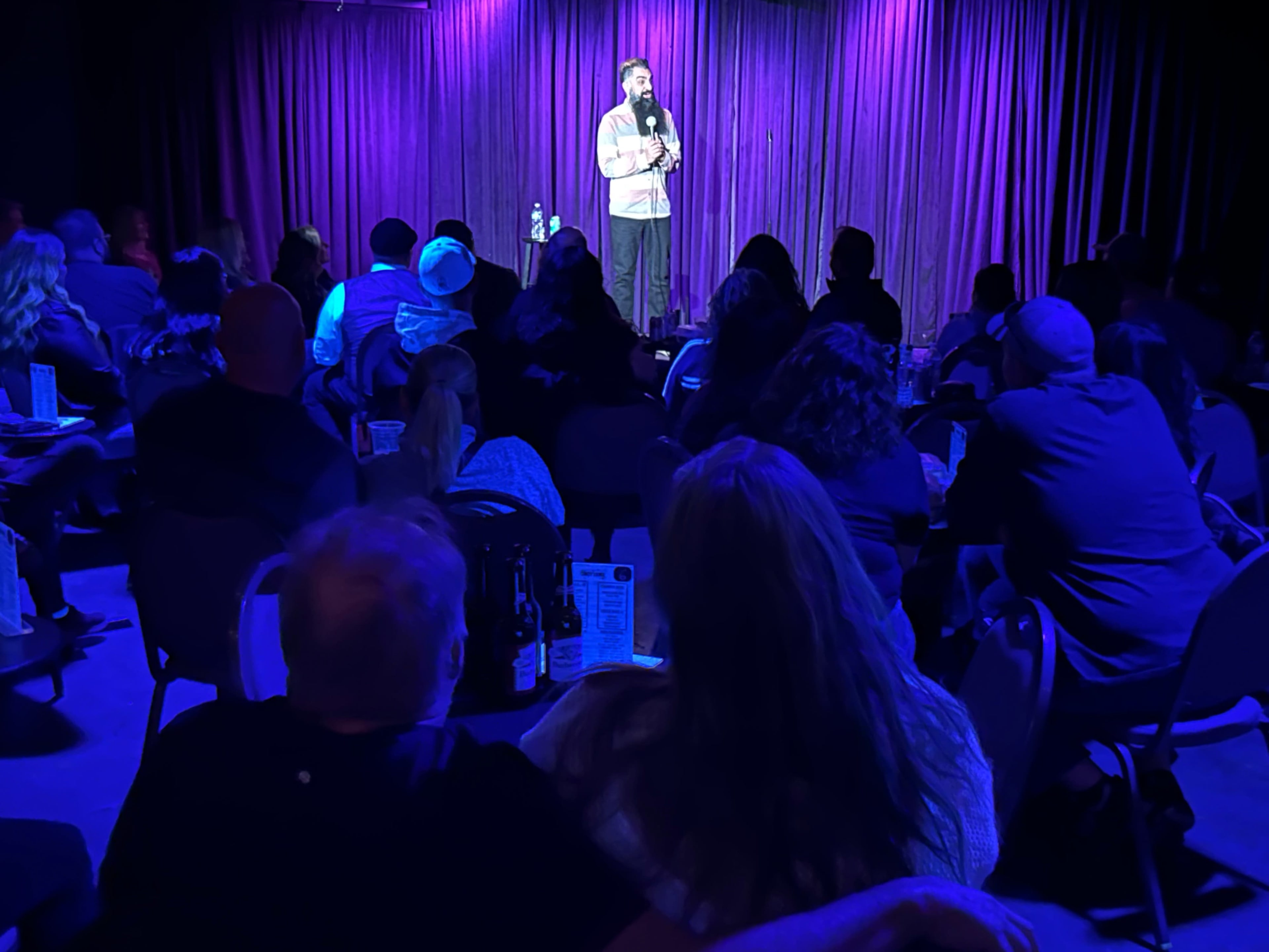 A comedian performs on stage in front of an audience seated at tables in a dimly lit venue with purple curtains.