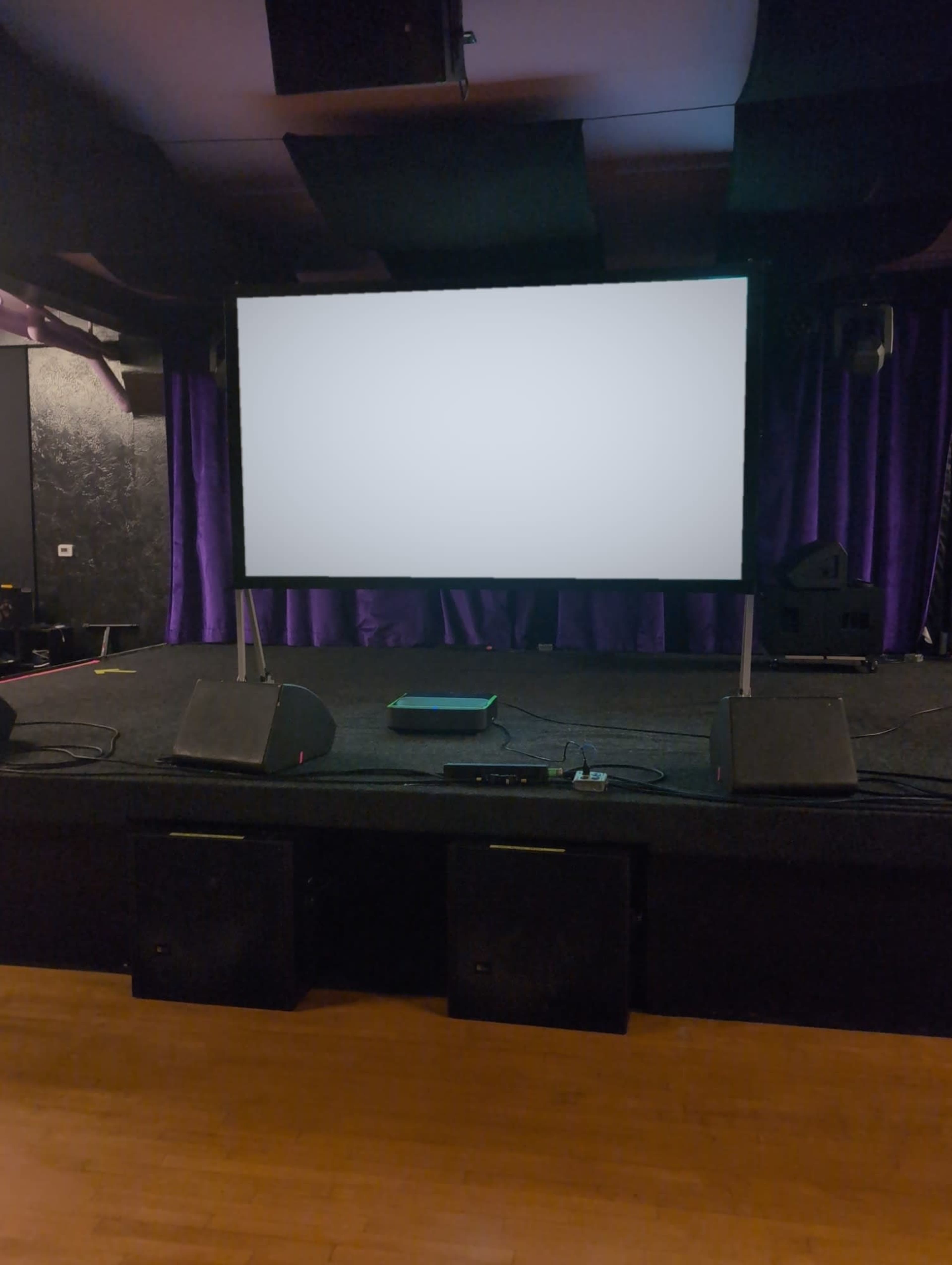 A blank projection screen stands on a stage surrounded by speakers and a dark backdrop with purple curtains.