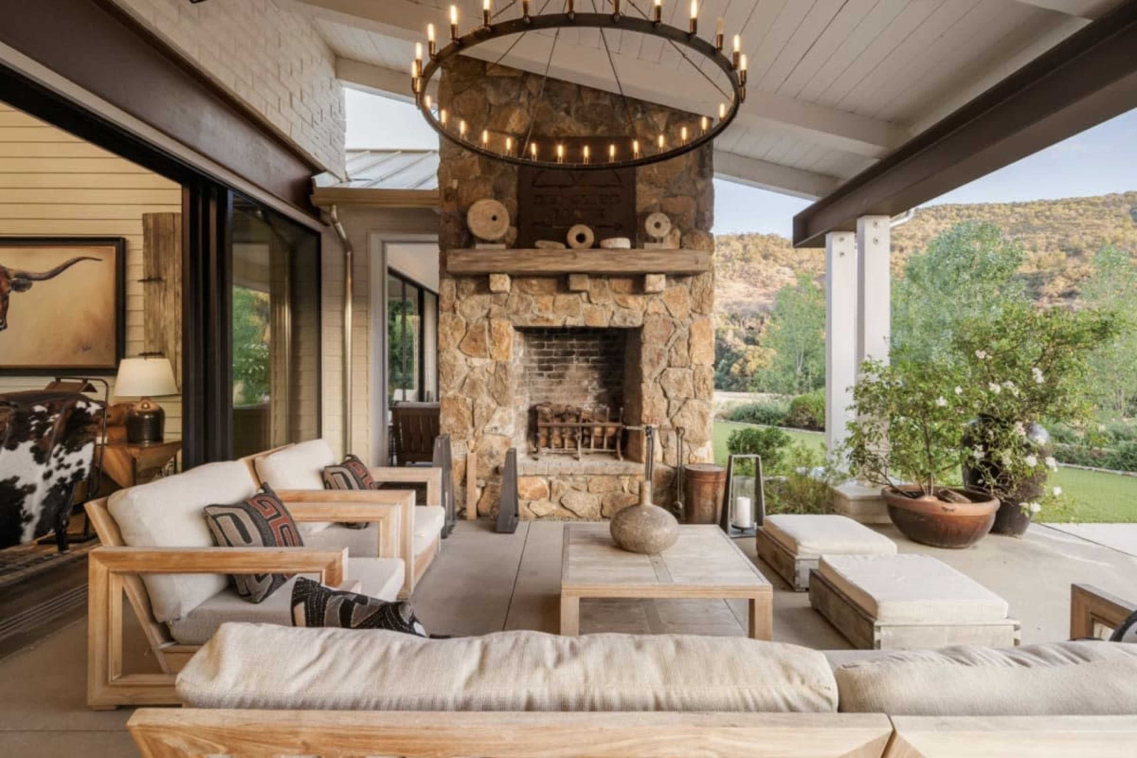 The image shows a spacious outdoor patio area featuring a stone fireplace, wooden furniture, and a large chandelier.