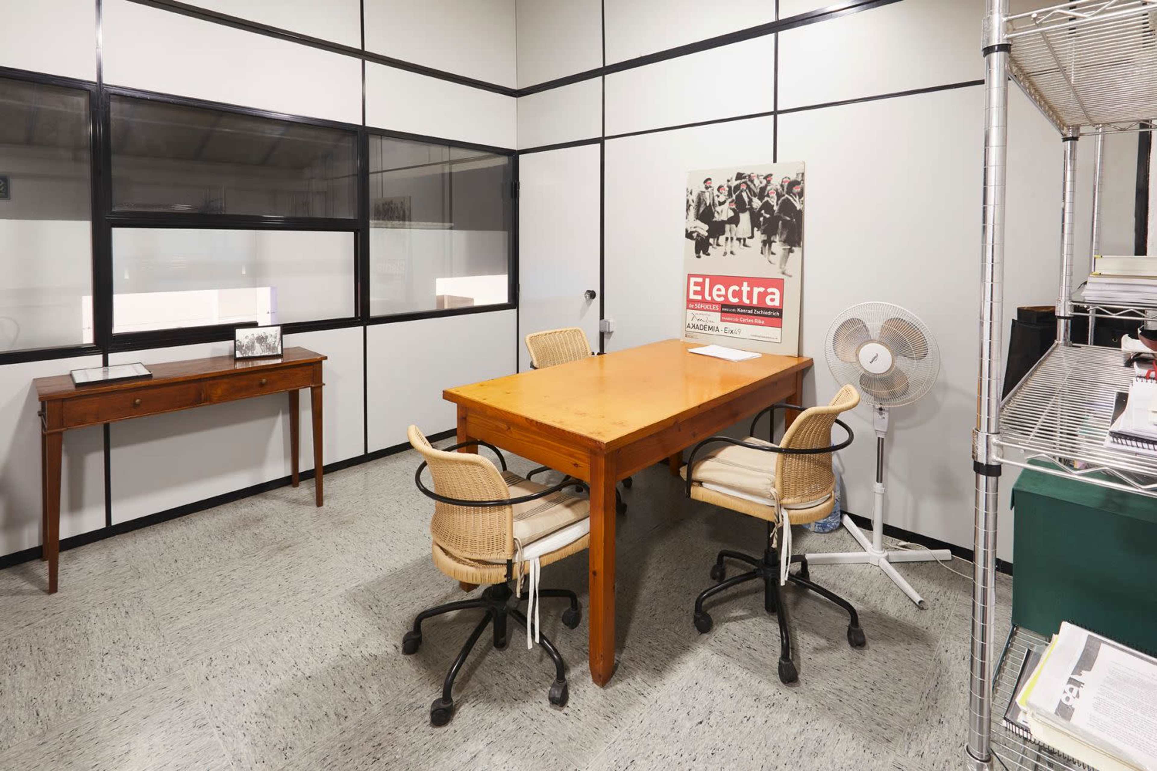 The image shows a small office with a wooden table surrounded by four chairs, a wall fan, and a framed photo on a side table.