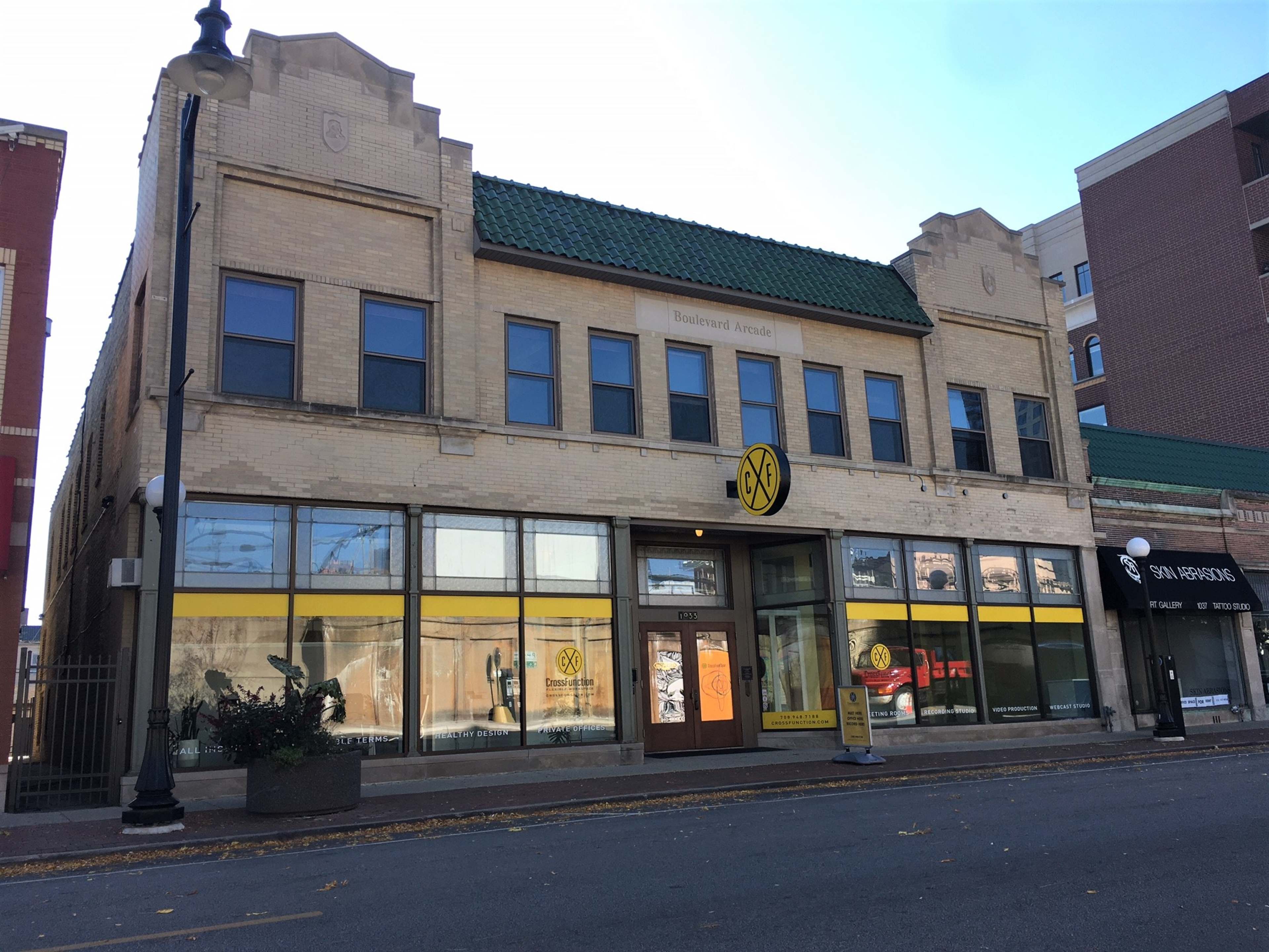 A brick commercial building with large front windows, a round black and yellow sign, and a green, tiled roof.