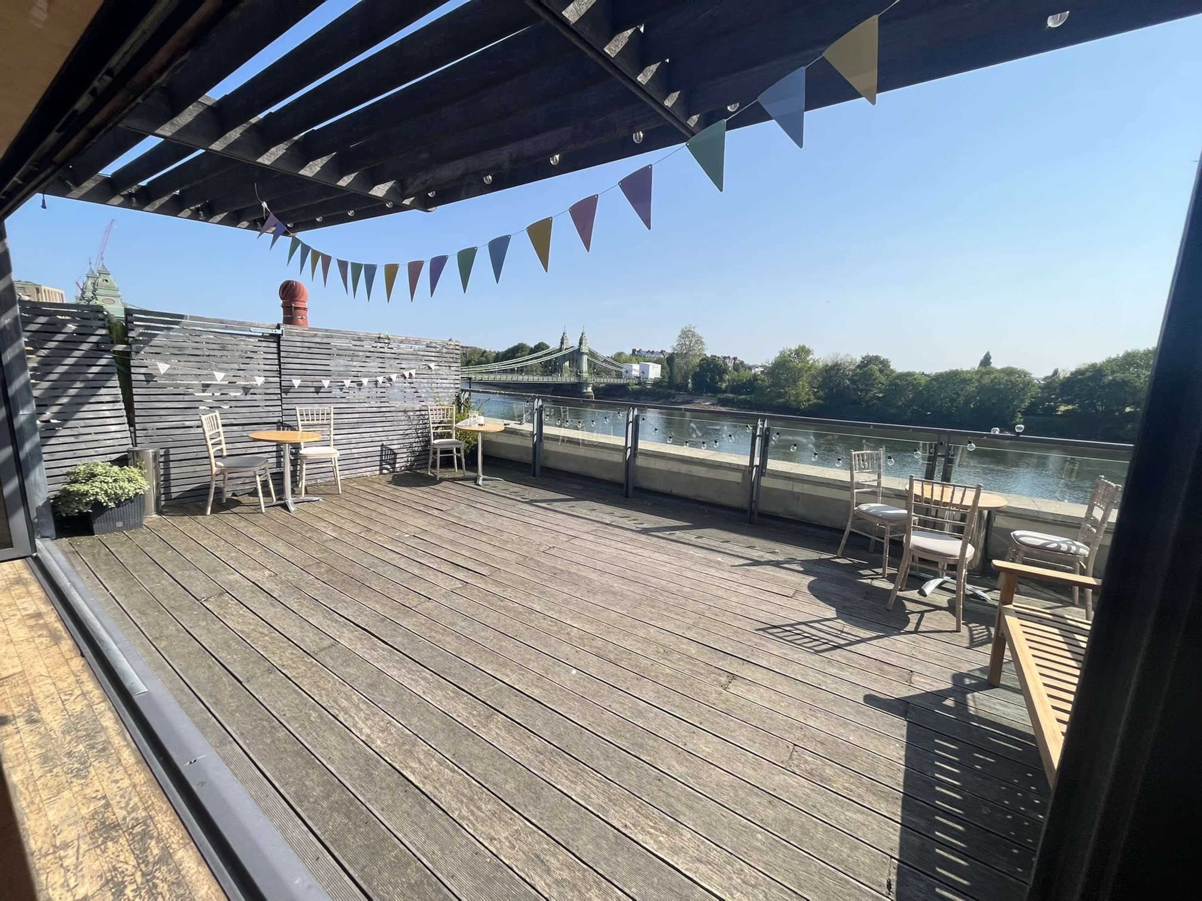 A wooden deck with chairs and tables overlooks a river, framed by colorful bunting and a clear blue sky.