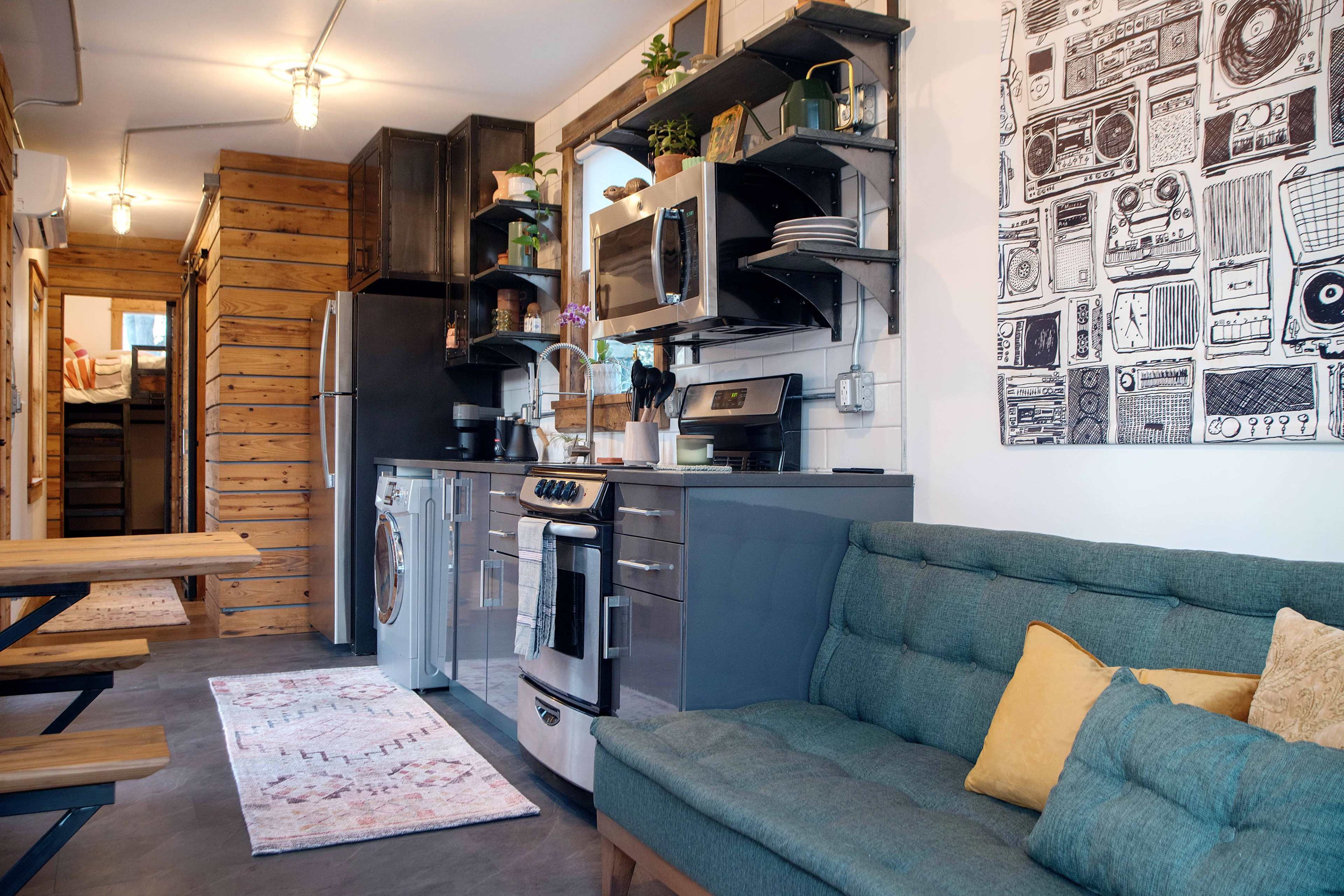 A compact kitchen area with modern appliances, wooden shelves, a sofa, and a decorative wall hanging.