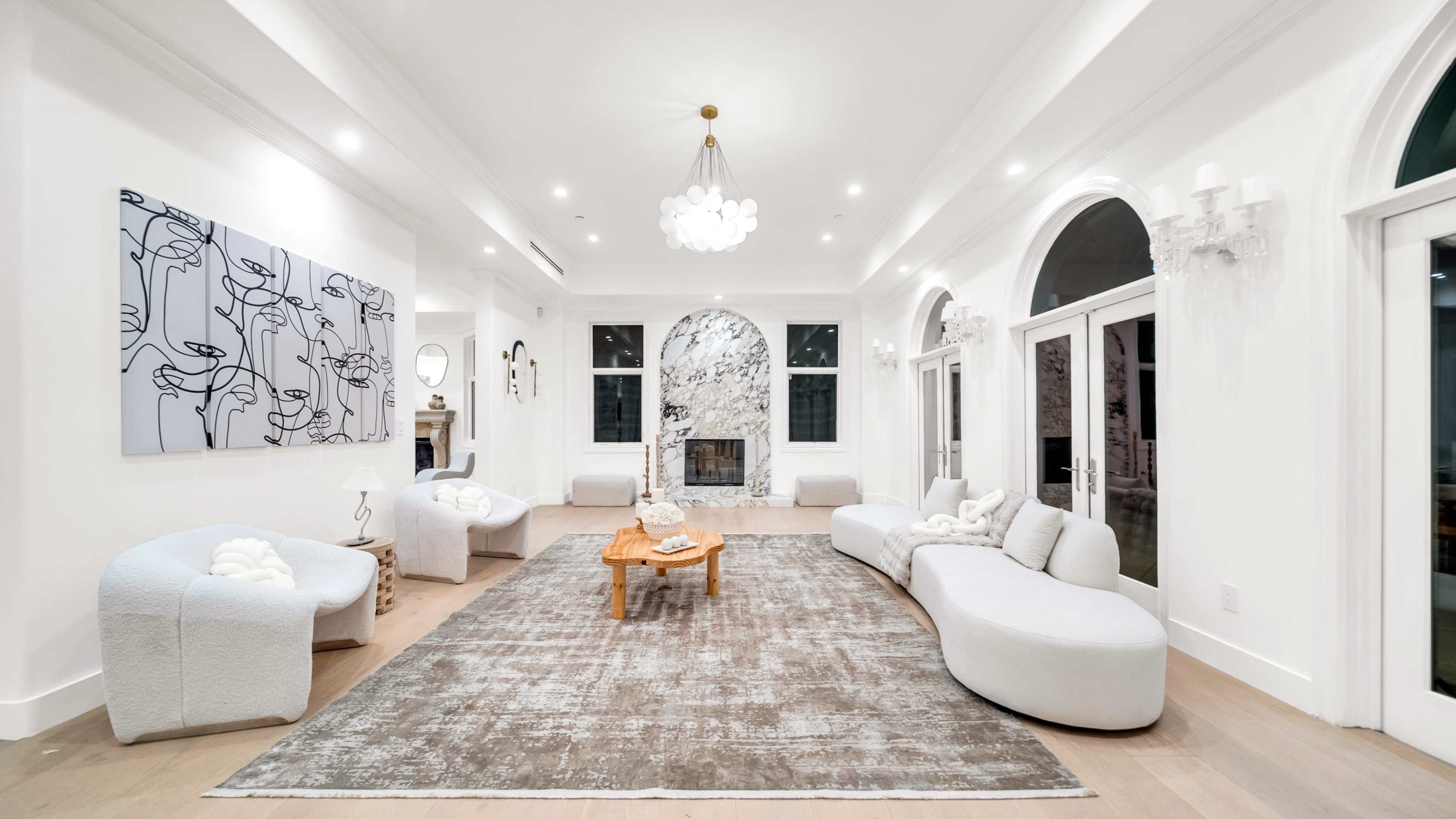 A spacious living room features modern furniture arranged around a central wooden table, with large windows and a decorative marble wall as a backdrop.