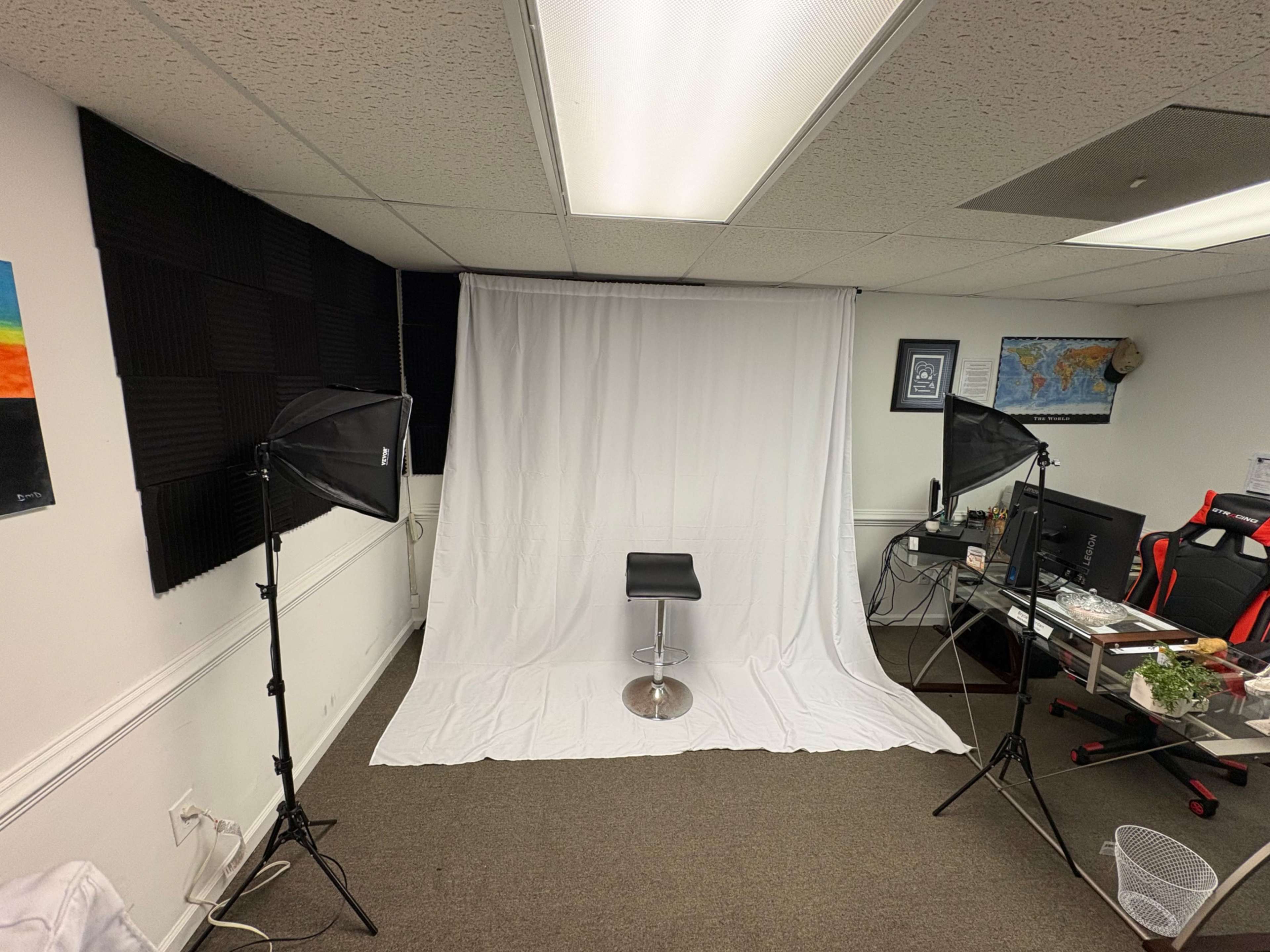 The image shows a photography studio setup with a white backdrop, a black stool, and lighting equipment, along with a desk and computer in the background.