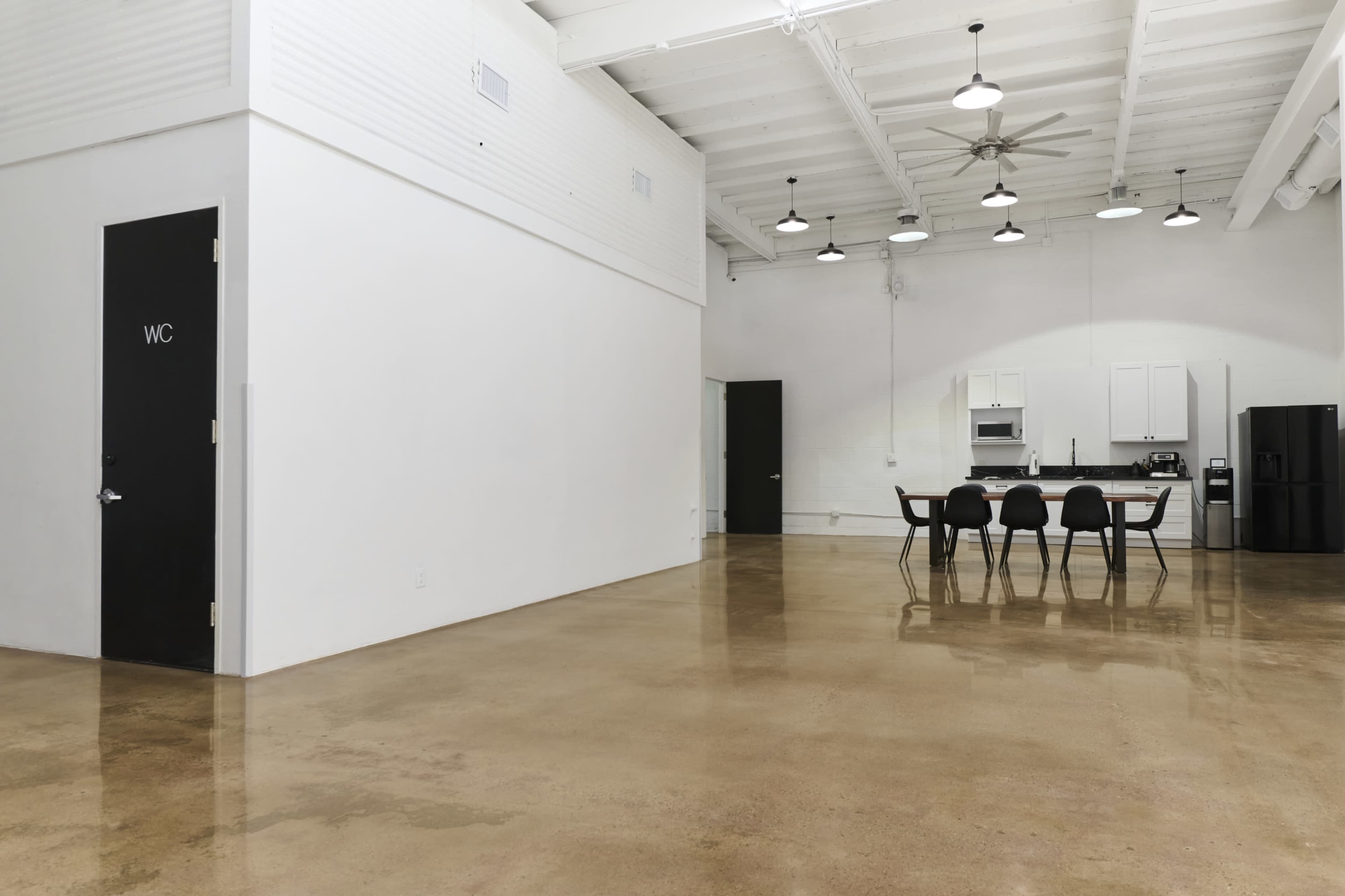 The image shows a spacious, modern interior with polished concrete floors, a white kitchen area, a dining table with black chairs, and a door labeled "WC" on the left.