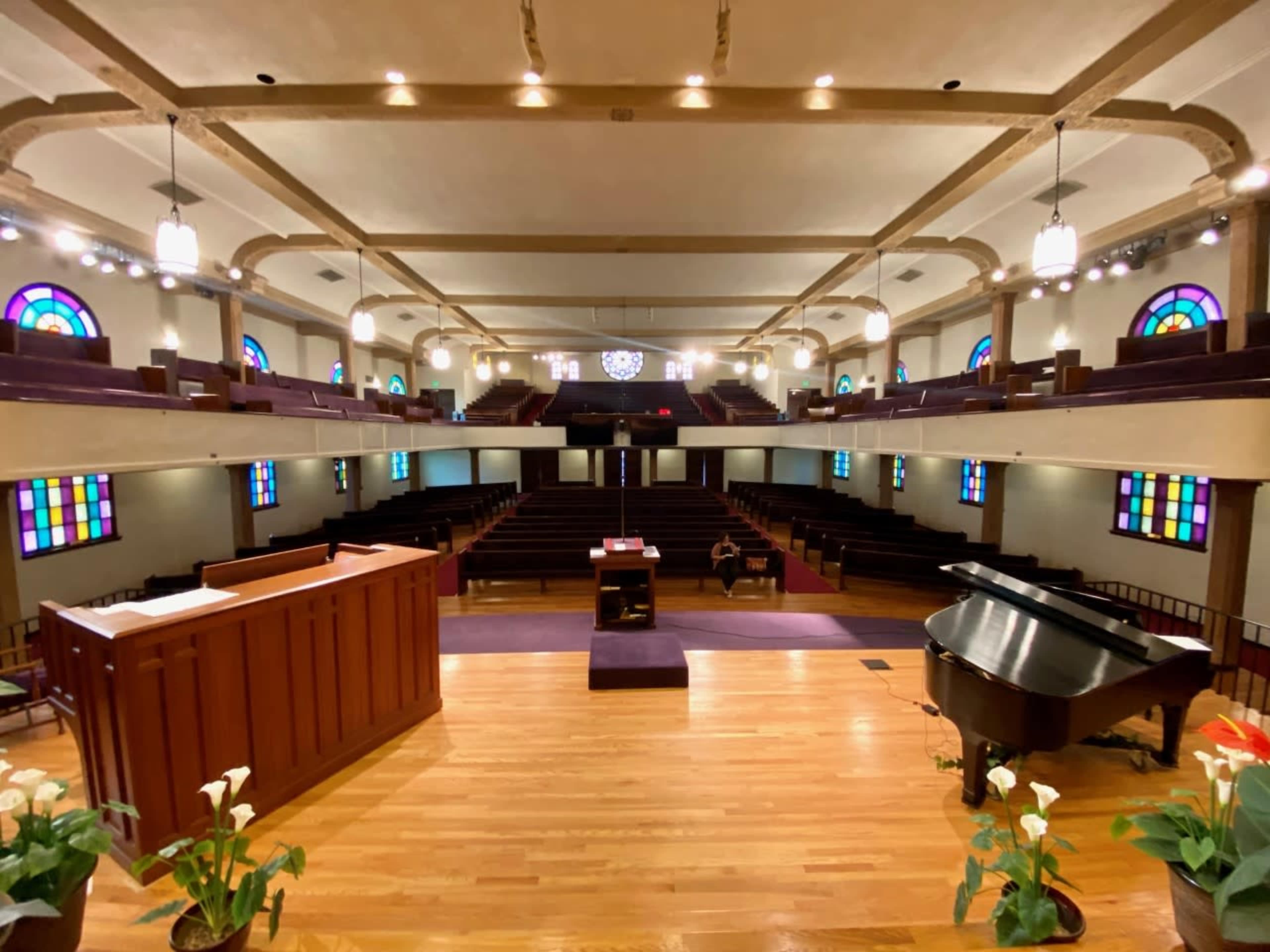 The interior of a church with a wooden pulpit, a grand piano, and colorful stained glass windows illuminating the empty pews.
