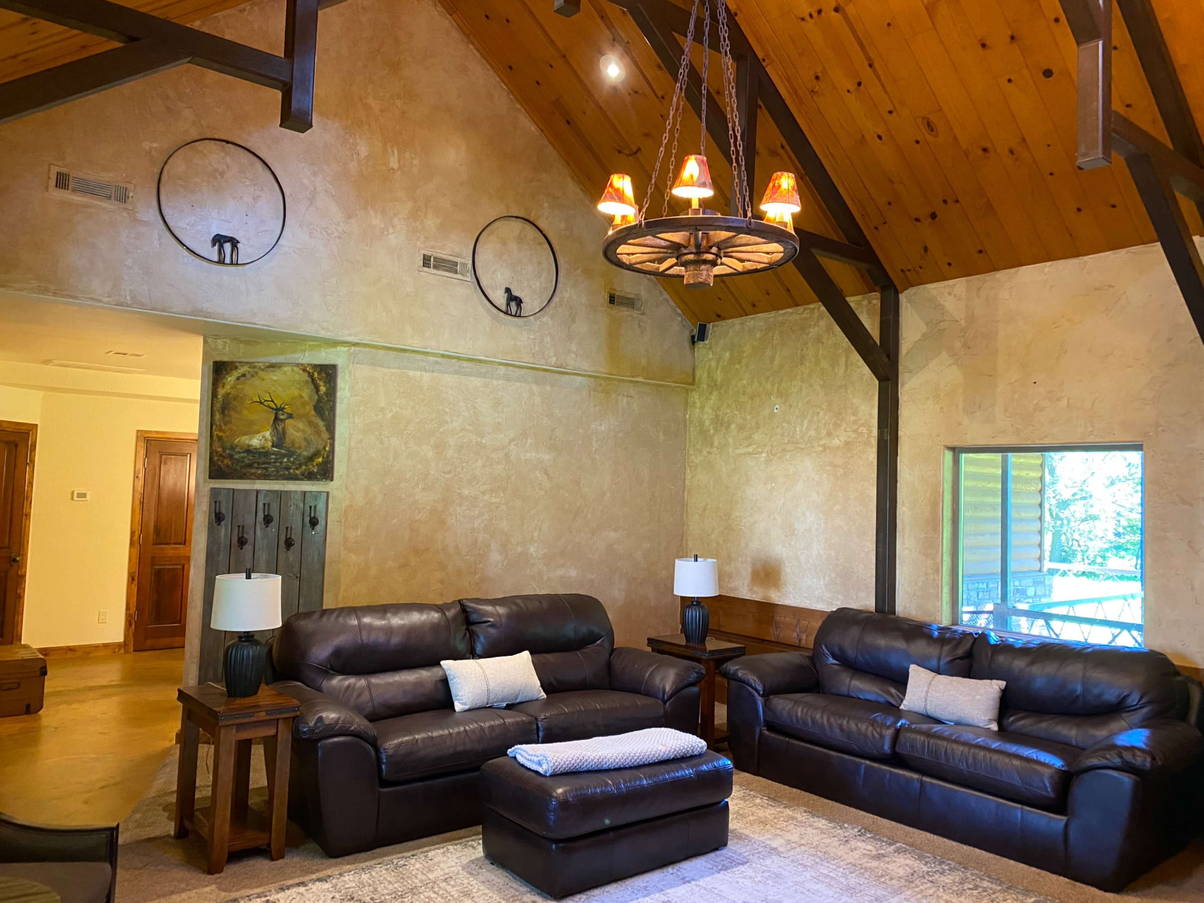 A spacious living room with two dark leather sofas, a central ottoman, and a large window, all set against a backdrop of wooden beams and a textured wall.