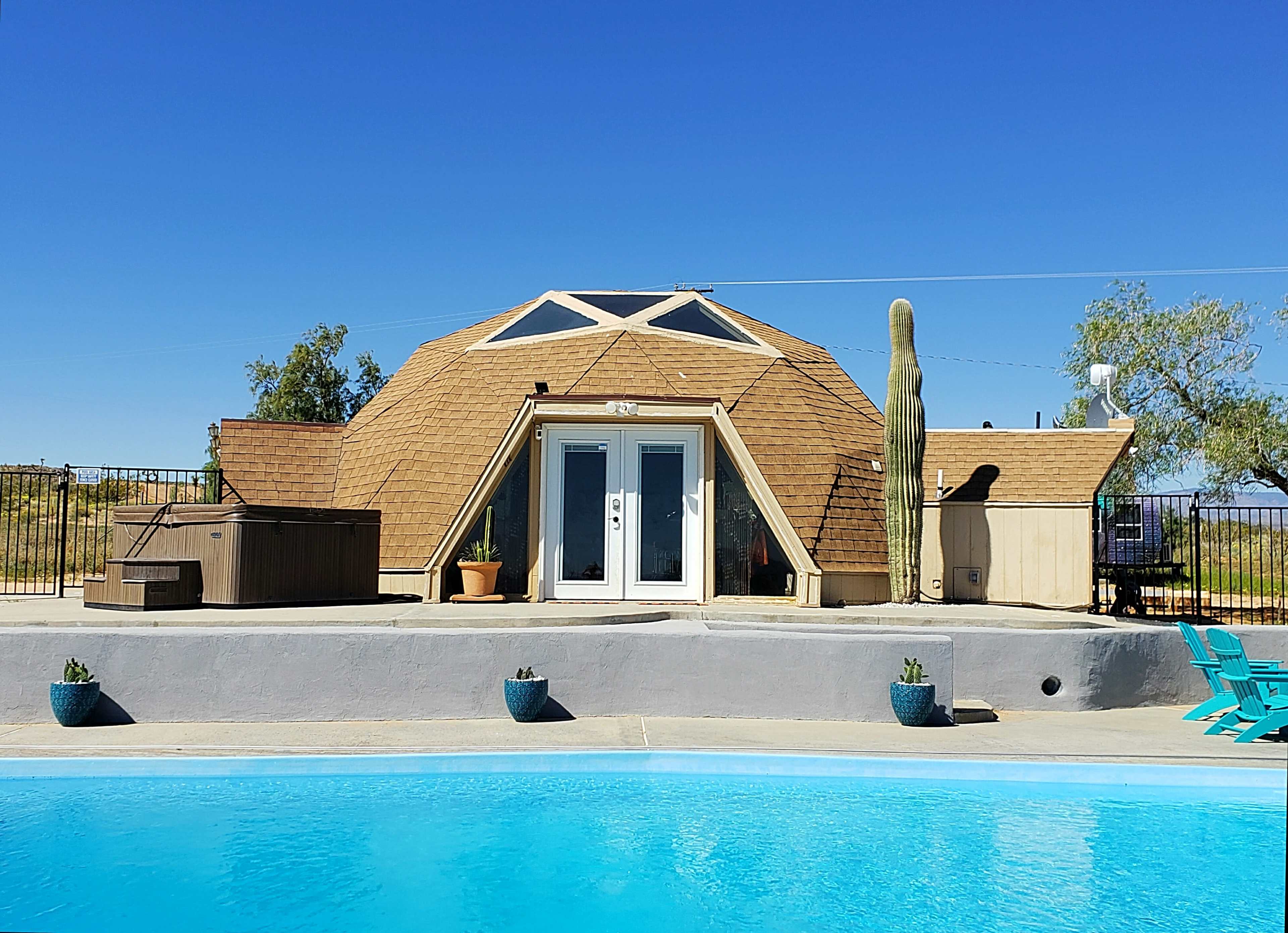 A geodesic dome house with a wooden exterior and large front doors stands beside a swimming pool, surrounded by cacti and desert landscaping.