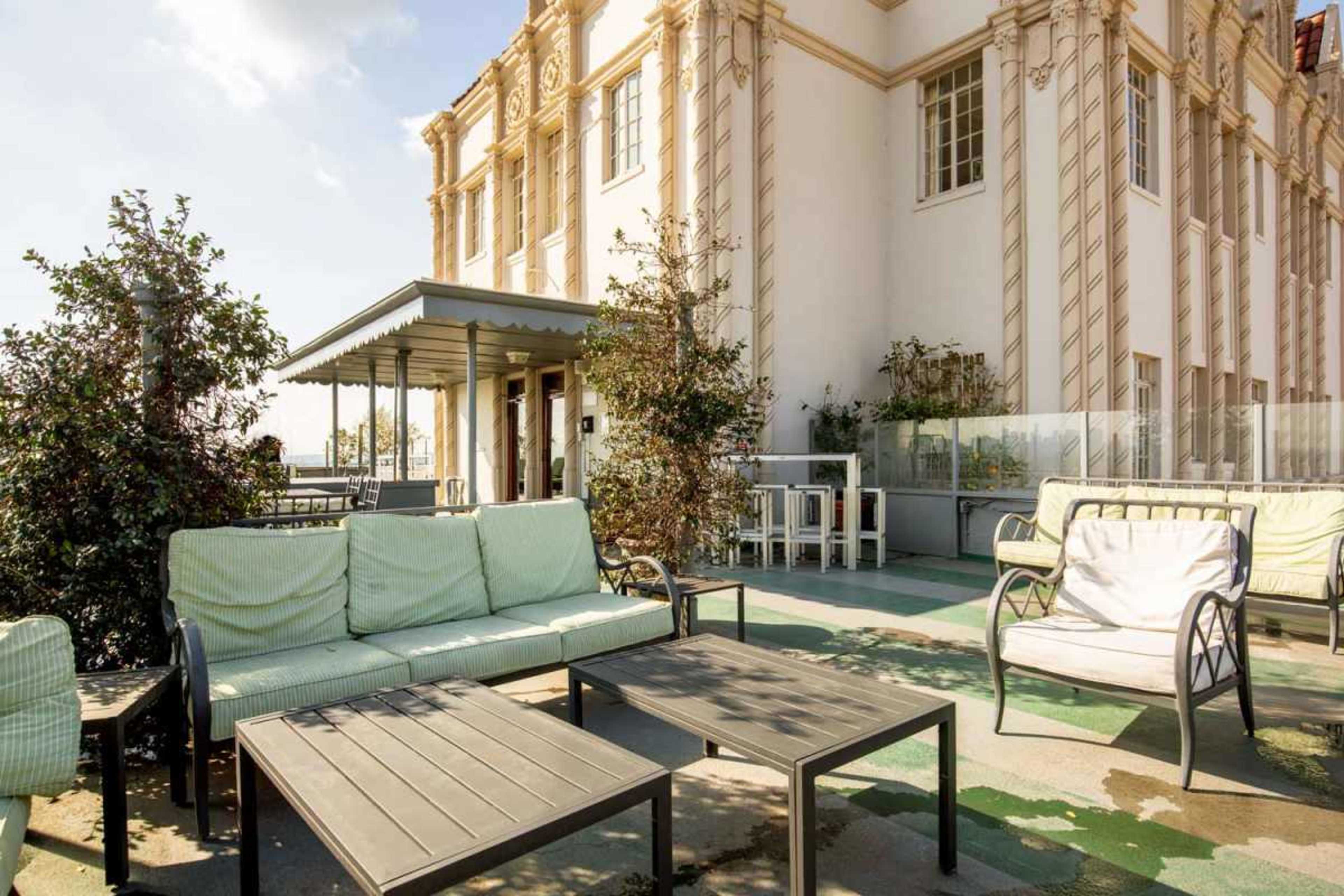 A rooftop terrace features metal tables and chairs surrounded by greenery and a building with ornate architectural details in the background.