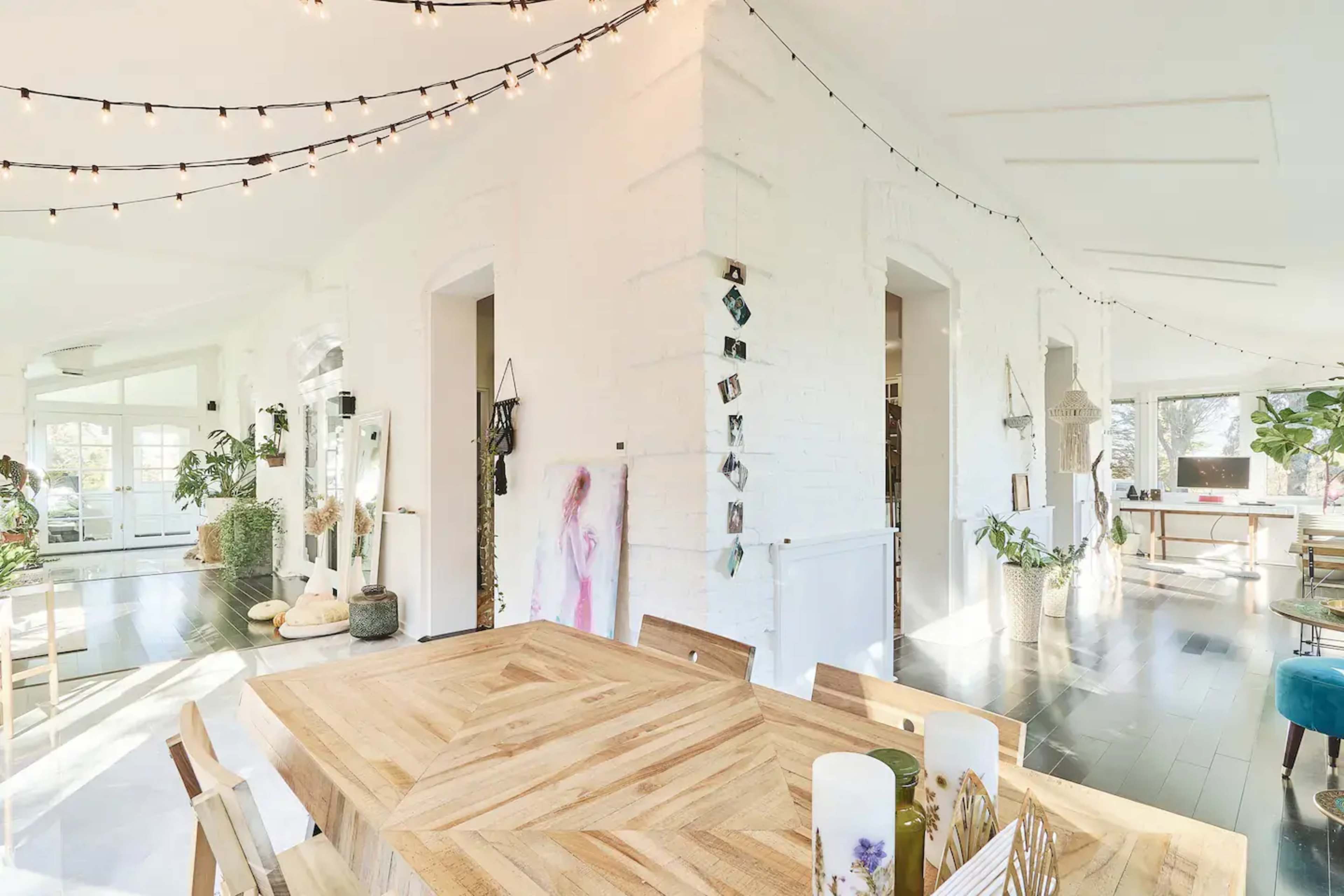 A well-lit, open dining area features a wooden table surrounded by chairs, with string lights and plants decorating the space.
