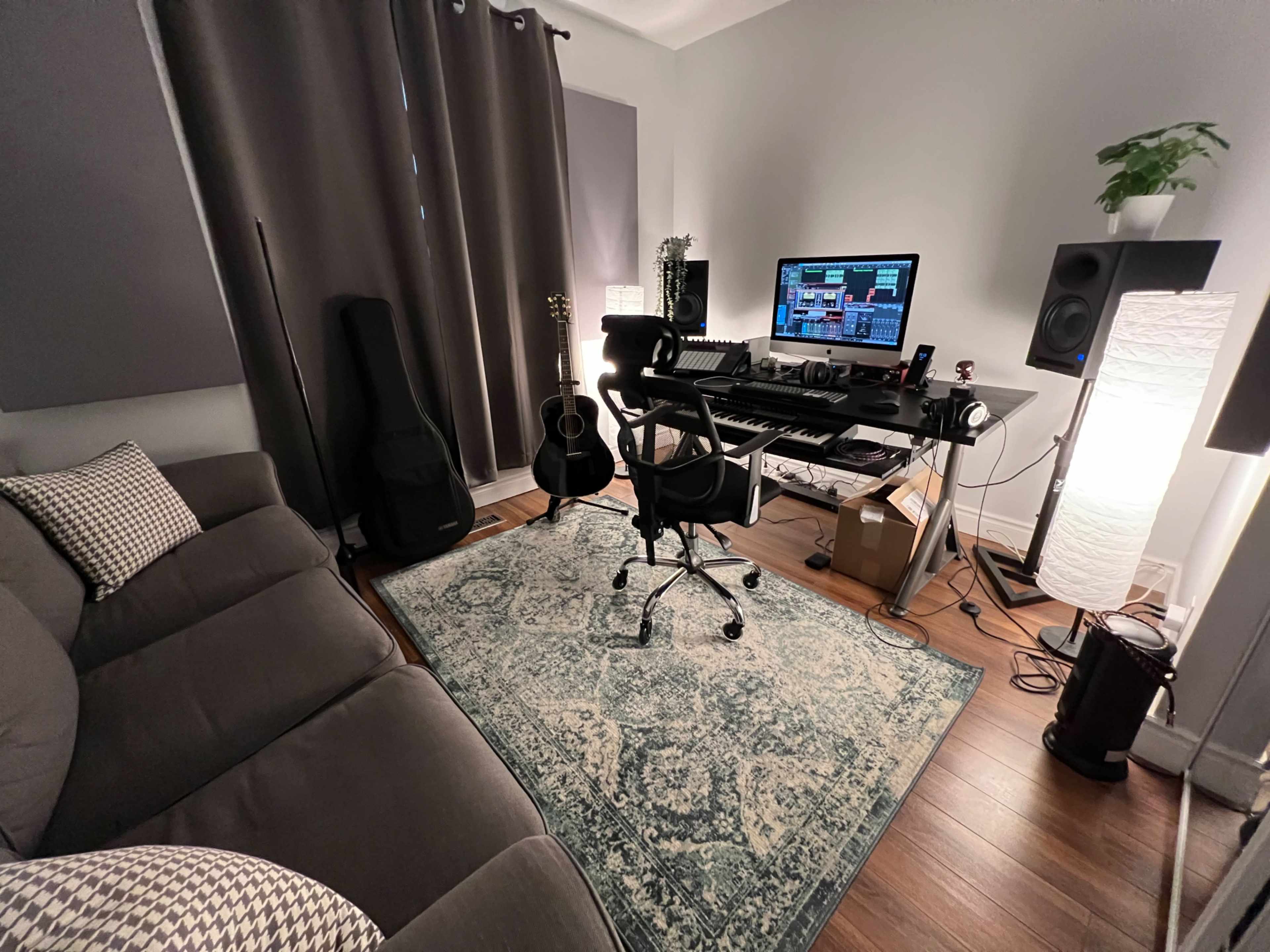 A home music studio featuring a desk with a computer and keyboard, a guitar leaning against a chair, and a cozy sofa with patterned cushions.