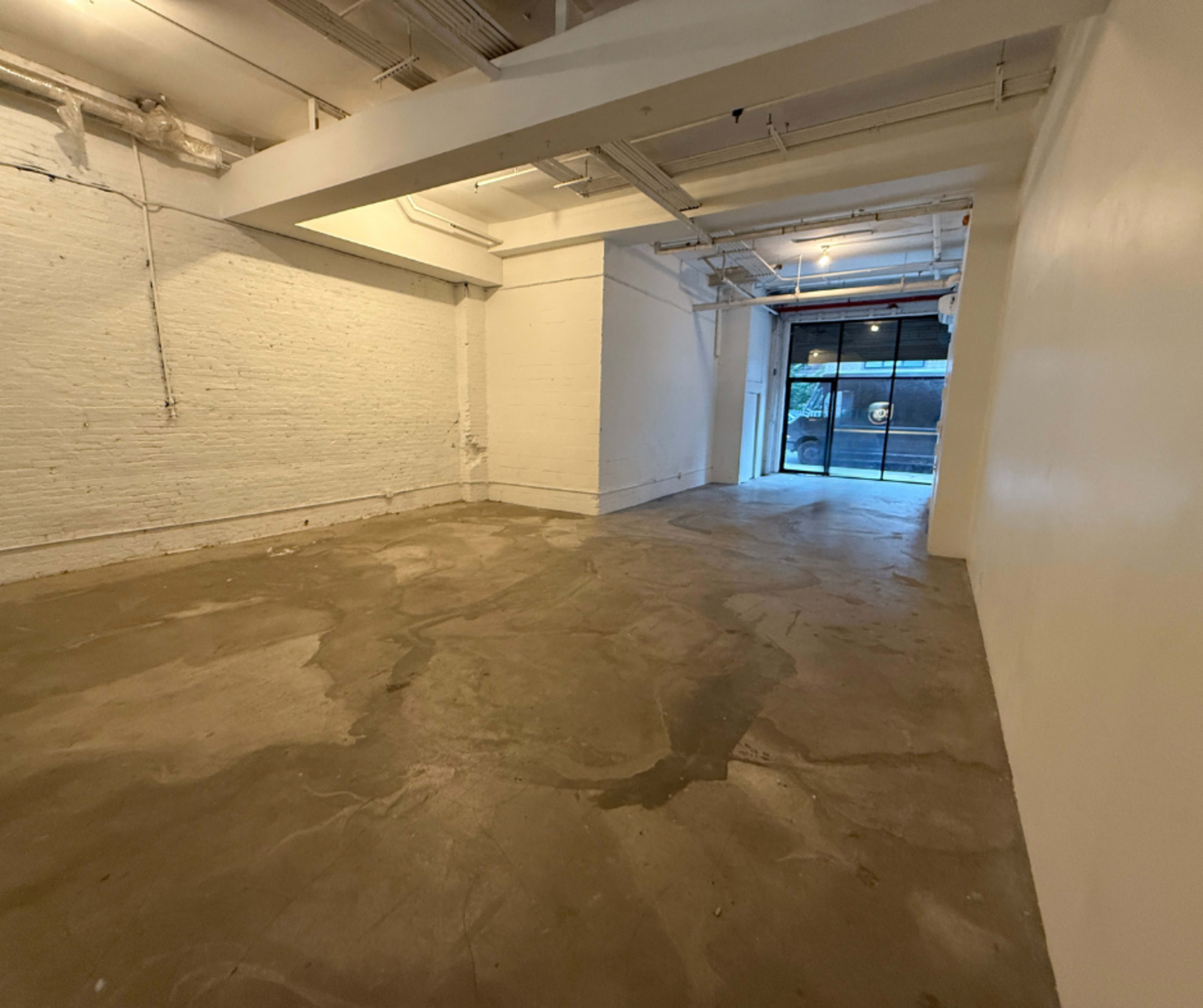 The image shows a spacious, empty room with concrete flooring and white walls, featuring a large window at the far end.