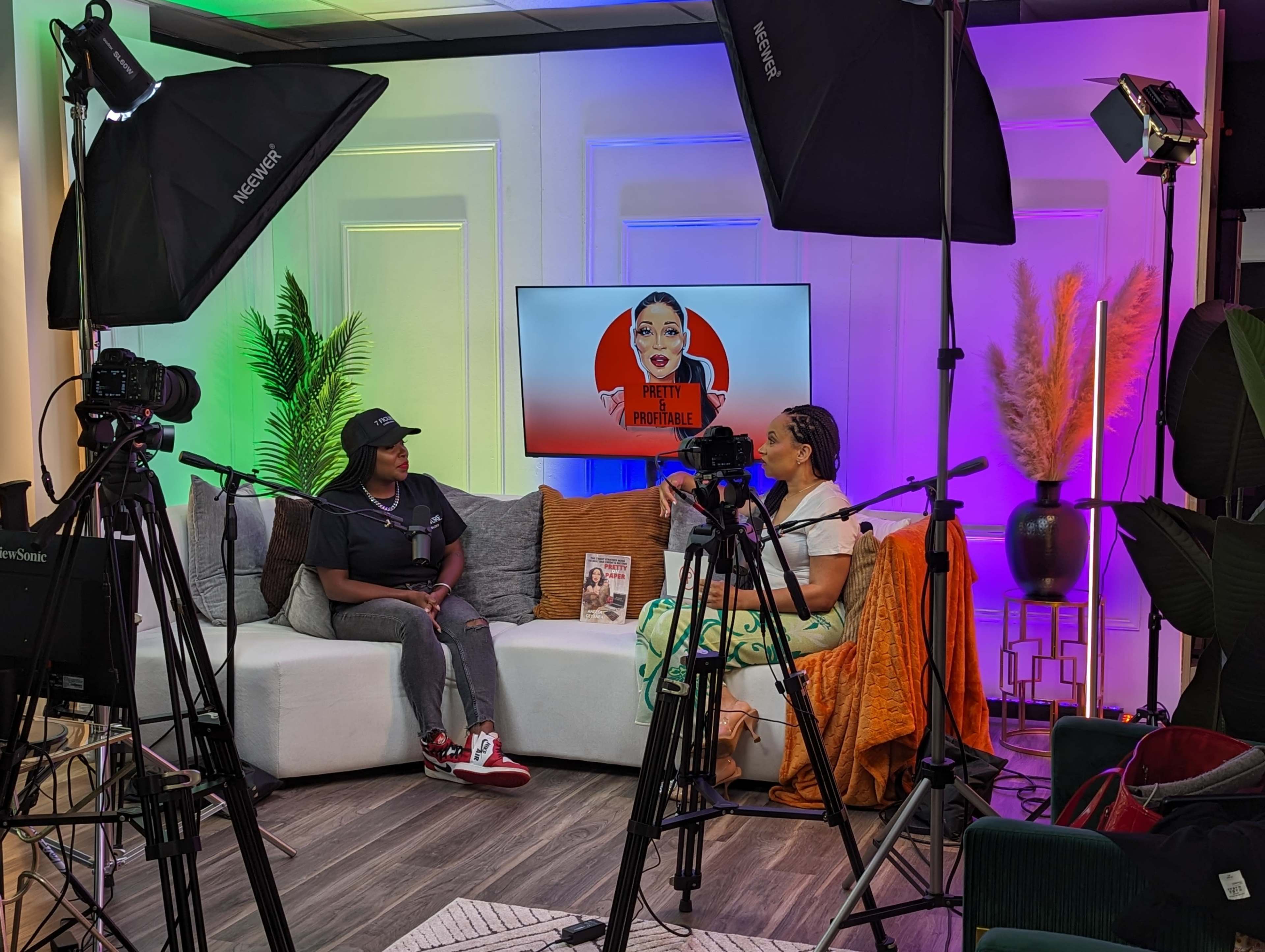 The scene features two individuals sitting on a couch in a well-lit studio set, surrounded by cameras, lighting equipment, and a screen displaying a promotional graphic.