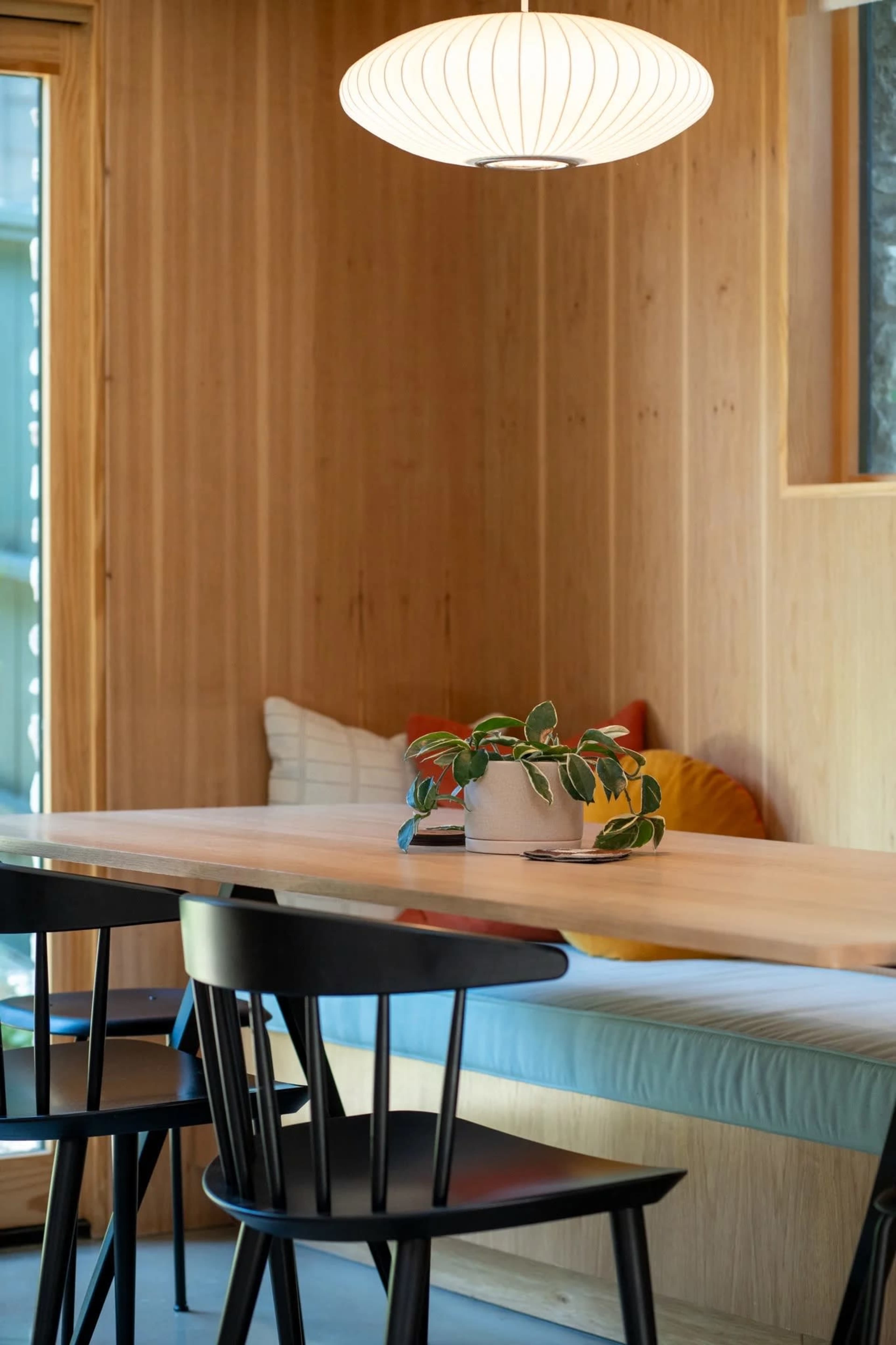 A wooden dining area features a light fixture above a table with a plant centerpiece and black chairs arranged around it.