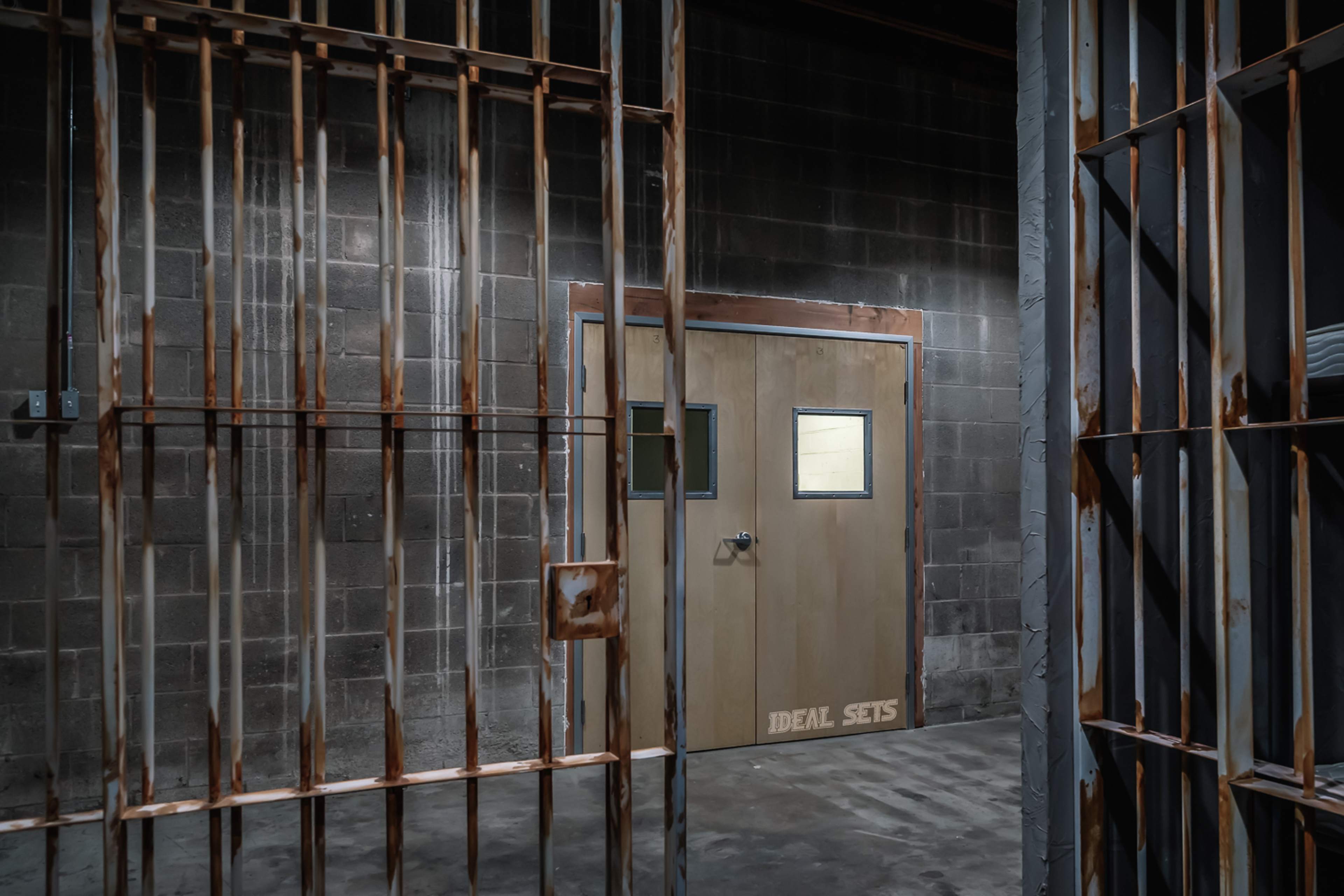 The image shows a dimly lit prison cell area with rusted bars and a solid door at the far end featuring small windows.