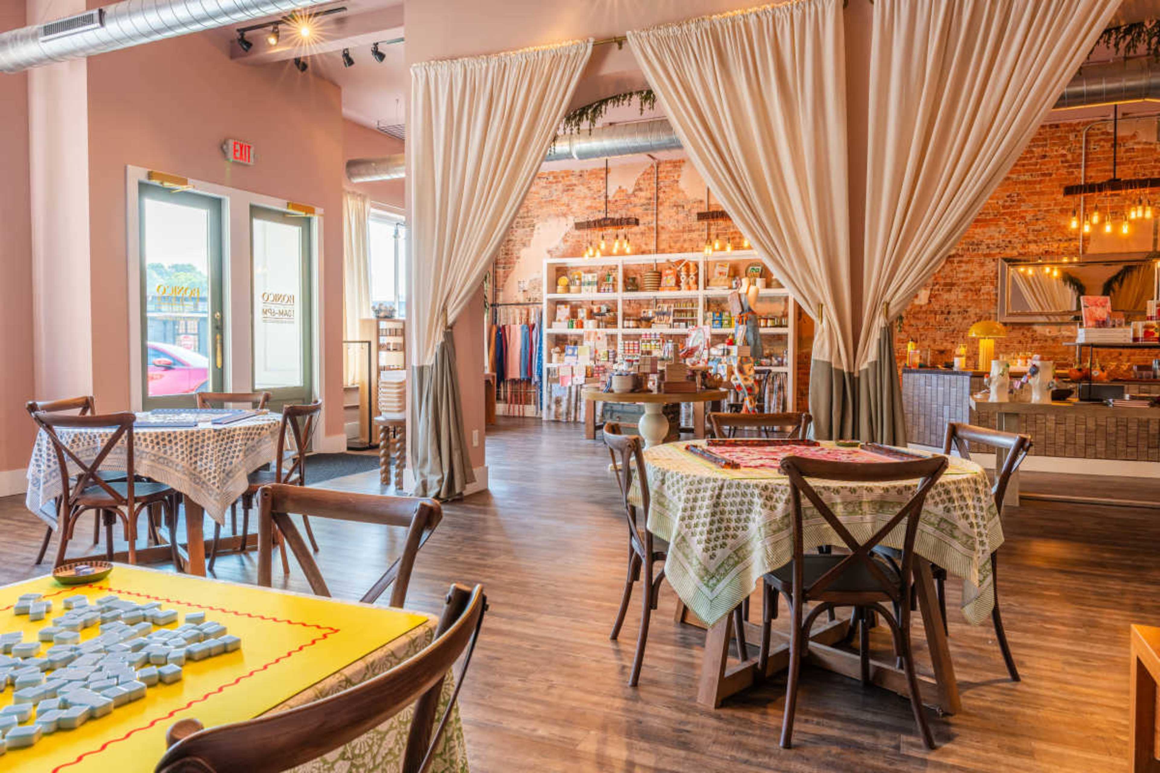 The image shows a spacious cafe with wooden tables, draped curtains, and a display of goods in the background.