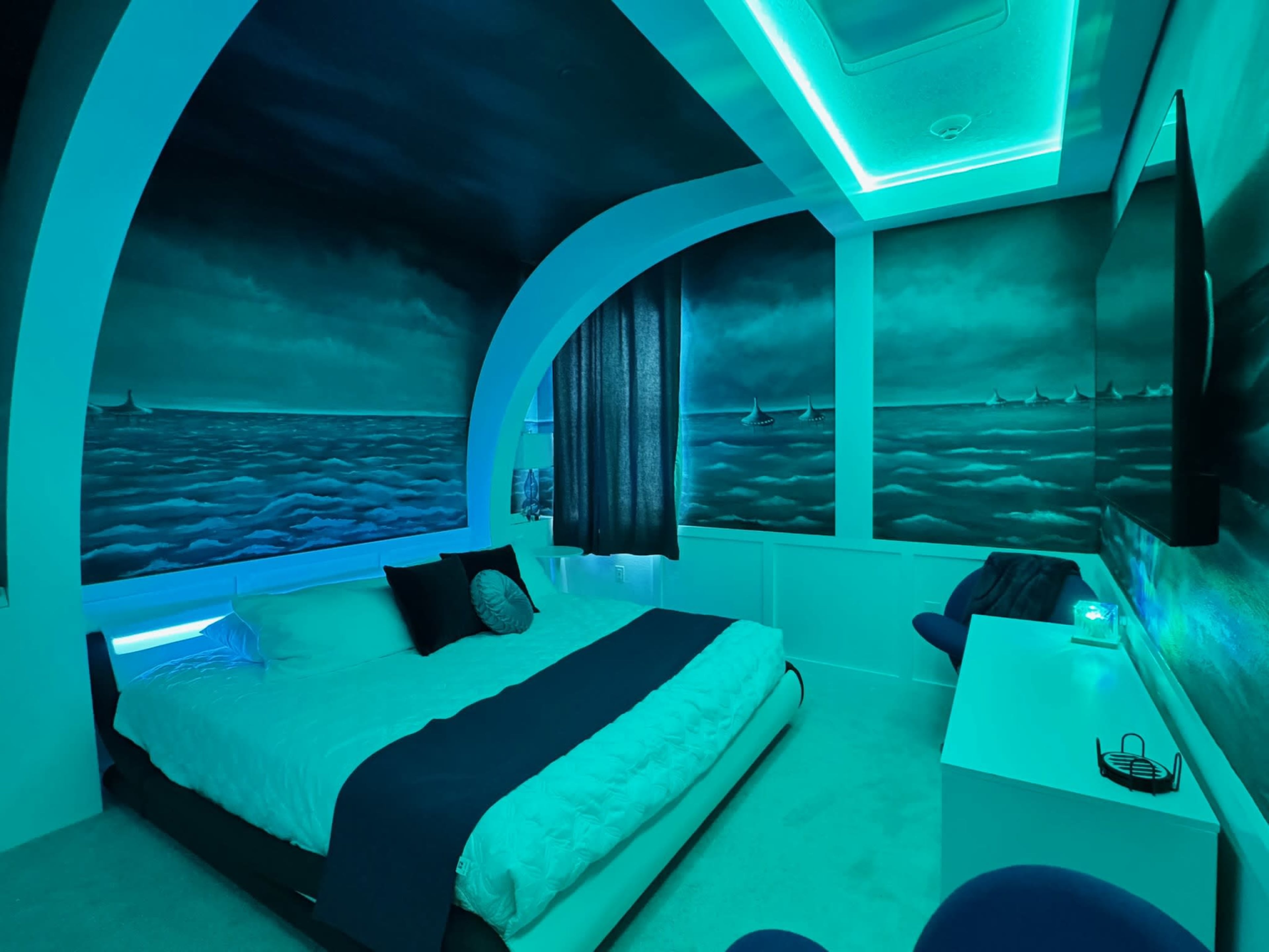 The image shows a modern bedroom with a large bed, ocean-themed murals on the walls, and blue LED lighting.