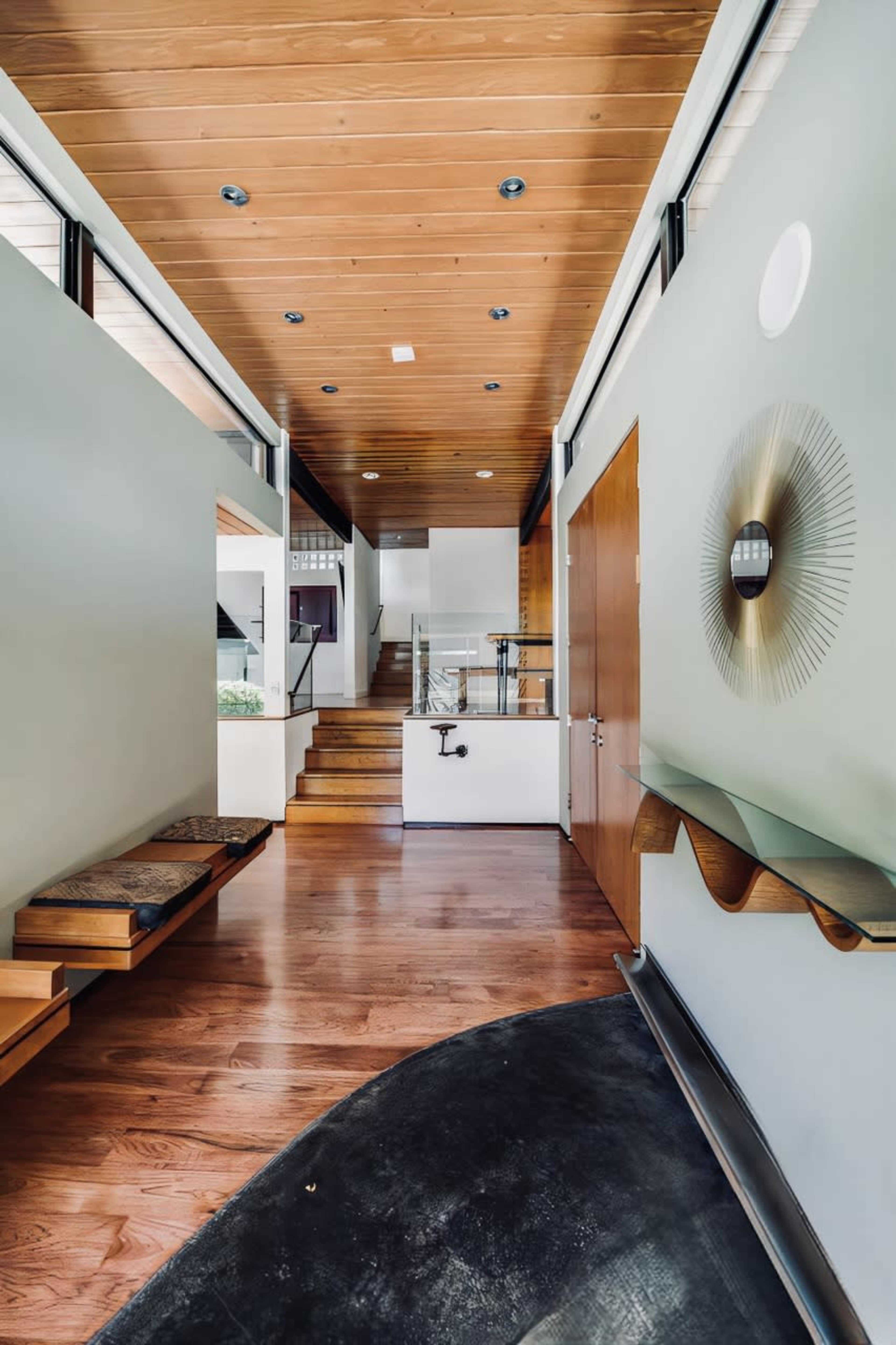 The image depicts a modern entryway with wooden ceilings, a sleek staircase, and large windows allowing natural light to flow in.