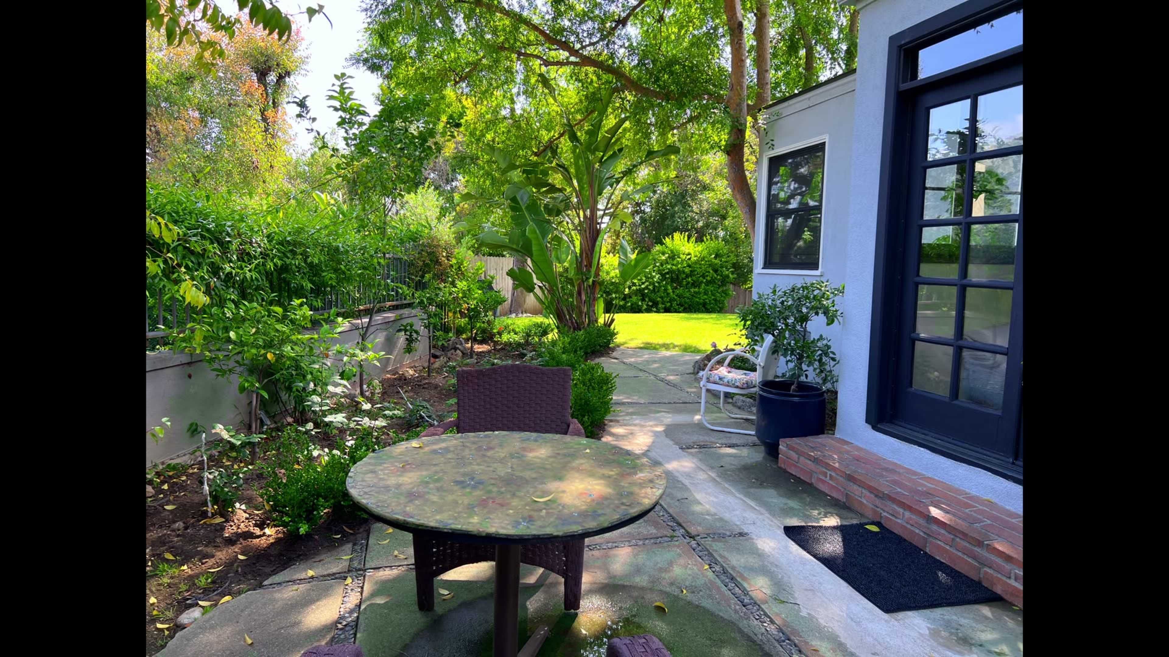 A circular table with chairs sits on a stone patio surrounded by lush greenery and a view of a lawn in a residential yard.