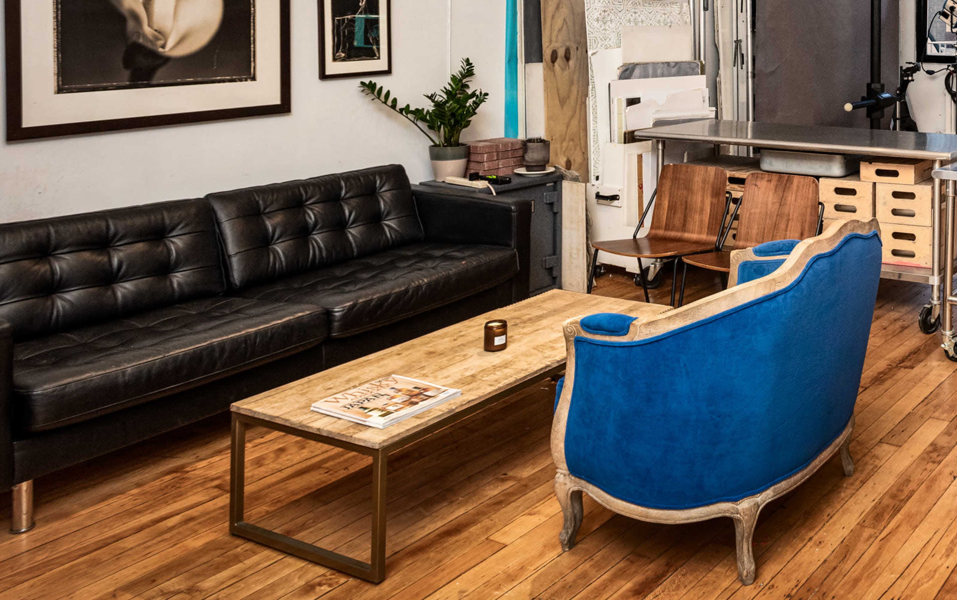 The image shows a living area with a black leather sofa, a wooden coffee table, two wooden chairs, and a blue upholstered chair, against a backdrop of wood flooring and framed artwork.