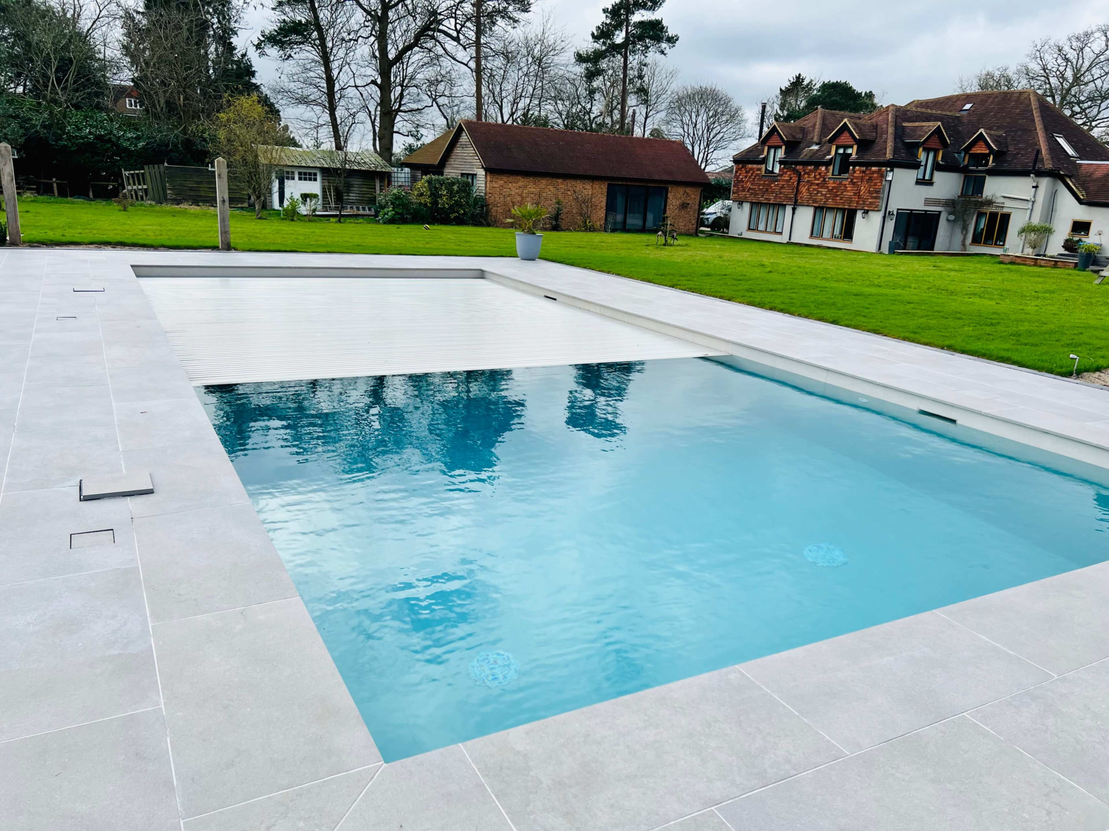A rectangular swimming pool surrounded by gray tiles and a grassy lawn, with a house and trees in the background.
