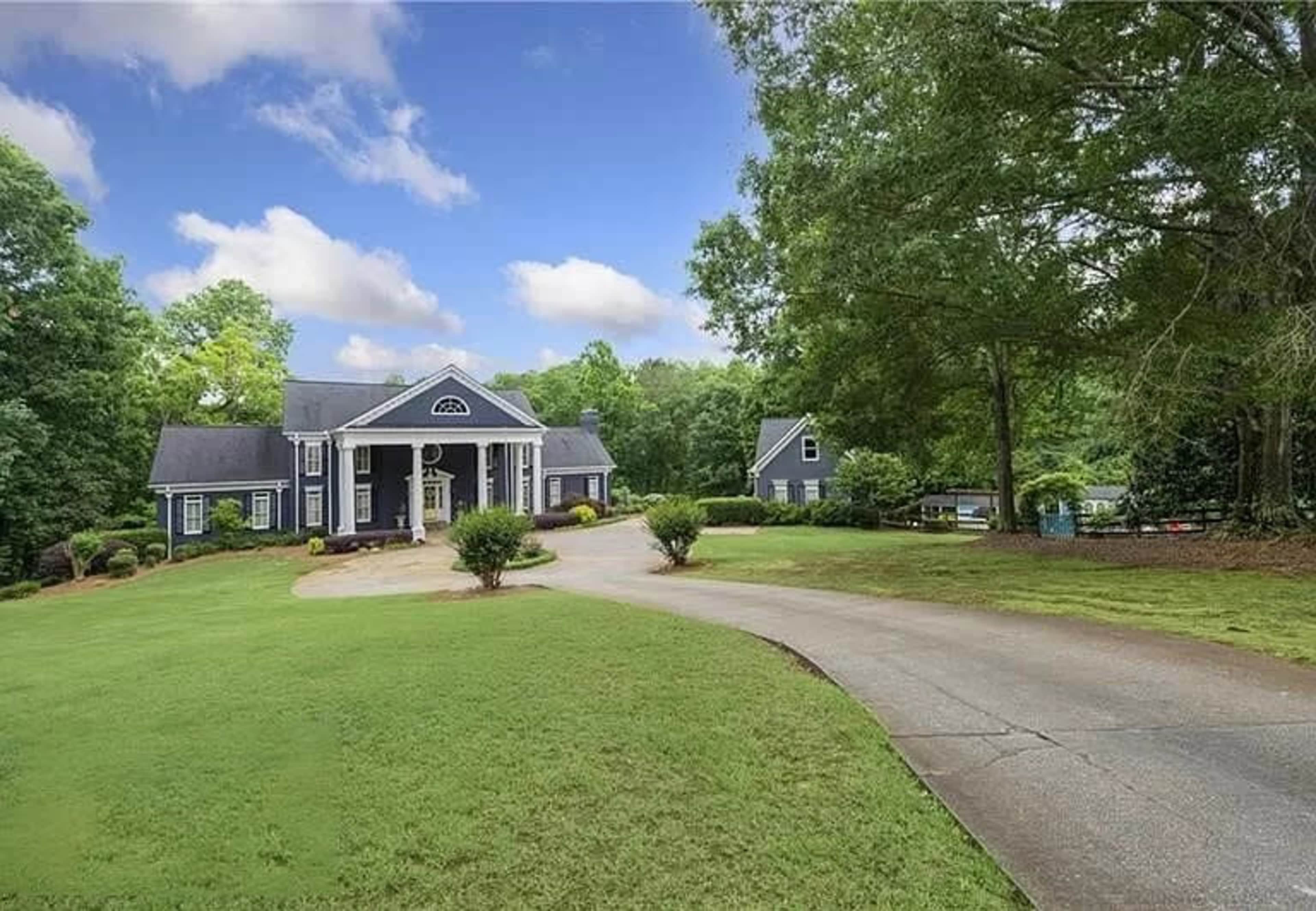 The image shows a large, two-story house with a circular driveway surrounded by greenery and trees.